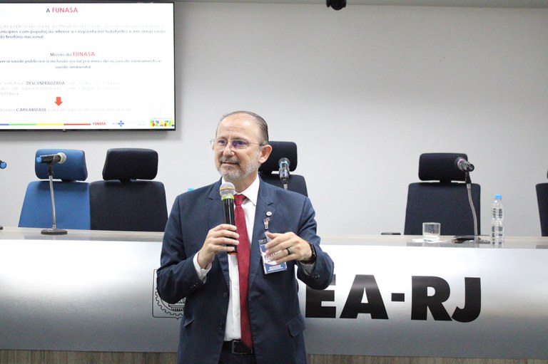 Alexandre Motta discursa em evento no CREA-RJ sobre fortalecimento do saneamento, segurando microfone, com painel institucional da Funasa ao fundo.