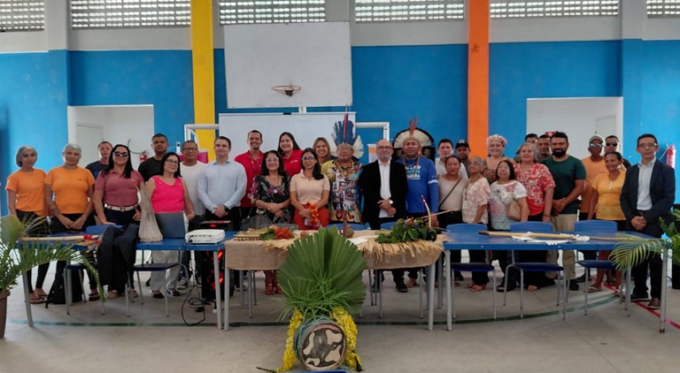 Grupo de pessoas reunidas em espaço comunitário, atrás de mesas com elementos culturais indígenas, em evento coletivo de integração e diálogo.