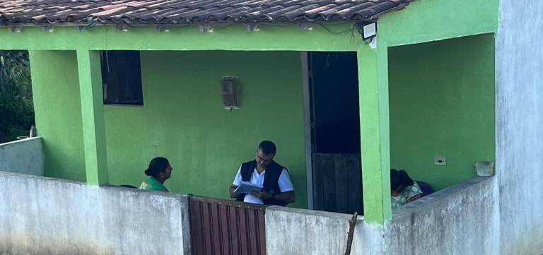 Servidor da Funasa registra informações em prancheta durante atendimento domiciliar a moradoras, em varanda de casa simples, com escuta ativa.