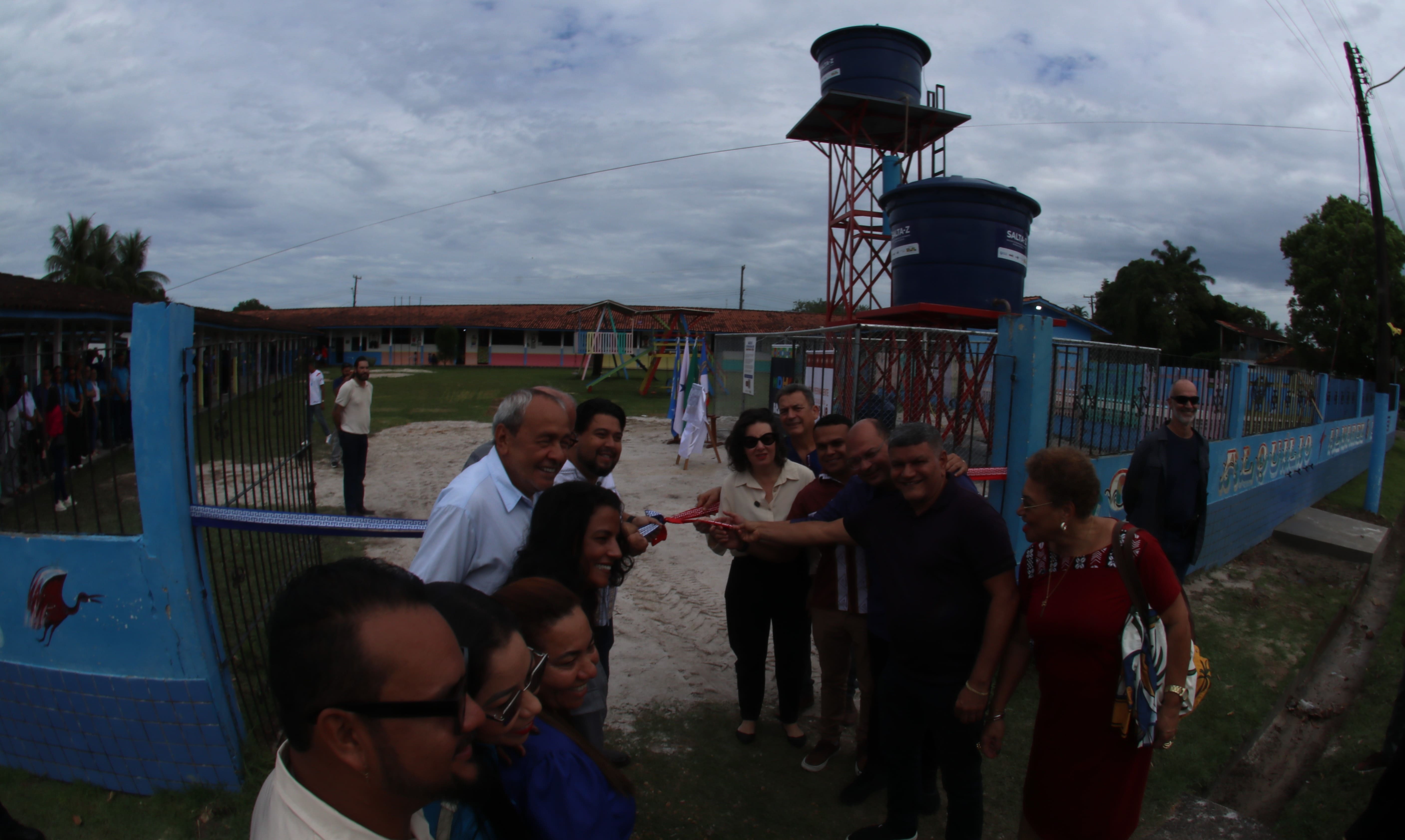Grupo de pessoas reunidas inaugurando sistema de água com fita vermelha em frente a uma escola.