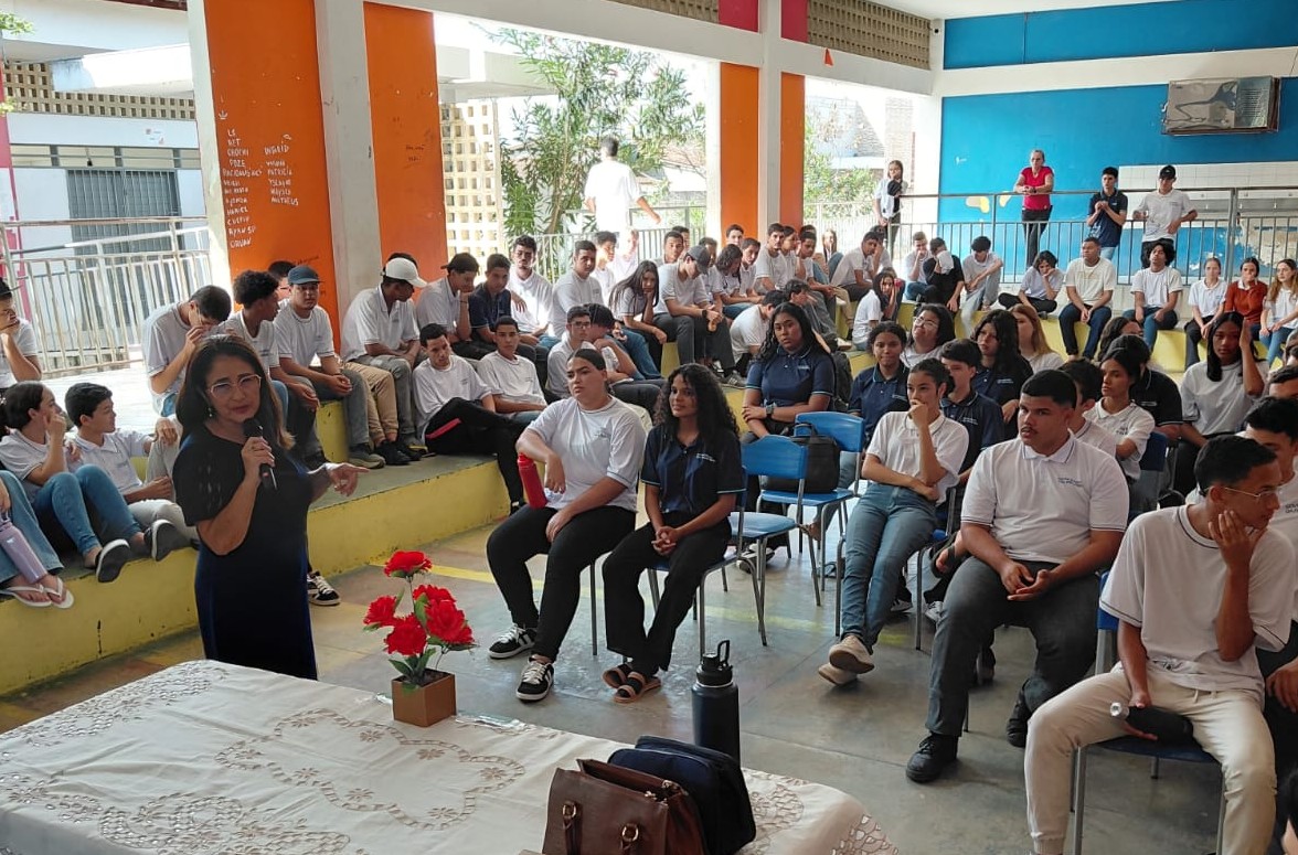 Diversas pessoas, alunos, reunidas em um pátio fechado, ouvindo uma palestra.