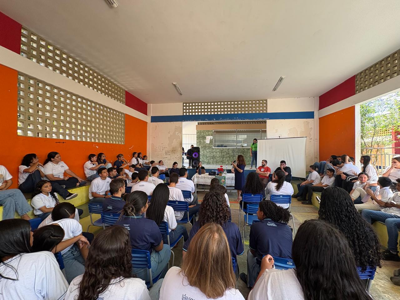 Do ângulo da plateia diversos alunos, reunidas em um pátio fechado, ouvindo uma palestra.
