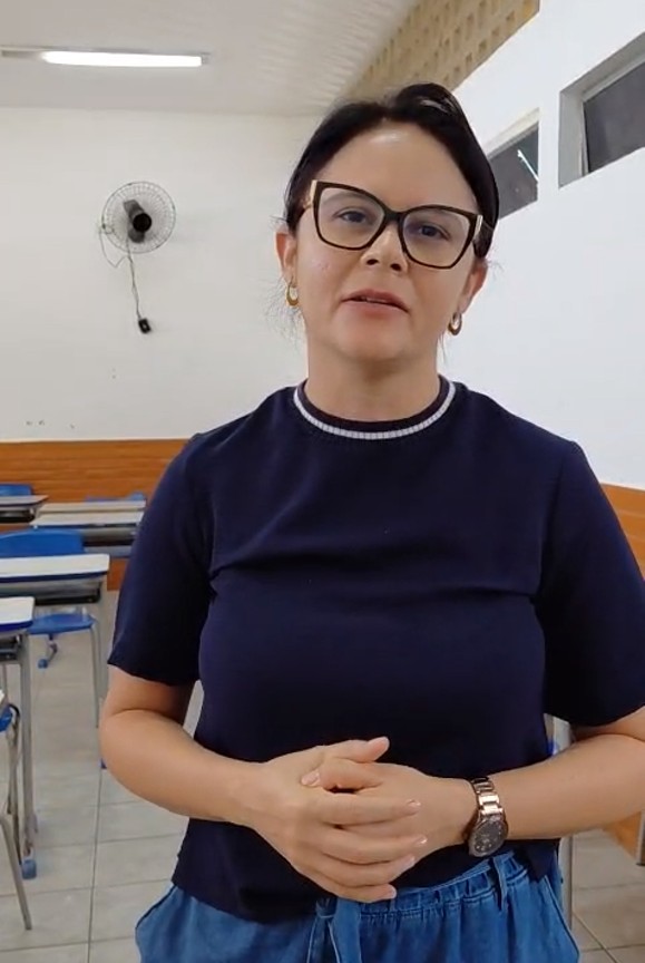 Retrato de uma professora de de óculos de armação preta, morena, meia idade, camiseta azul marinho.