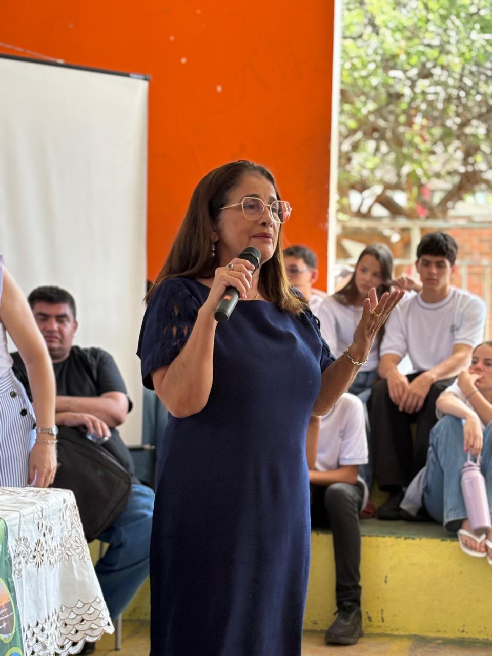 Mulher palestrando em sala com fundo laranja. Diversas crianças observam.