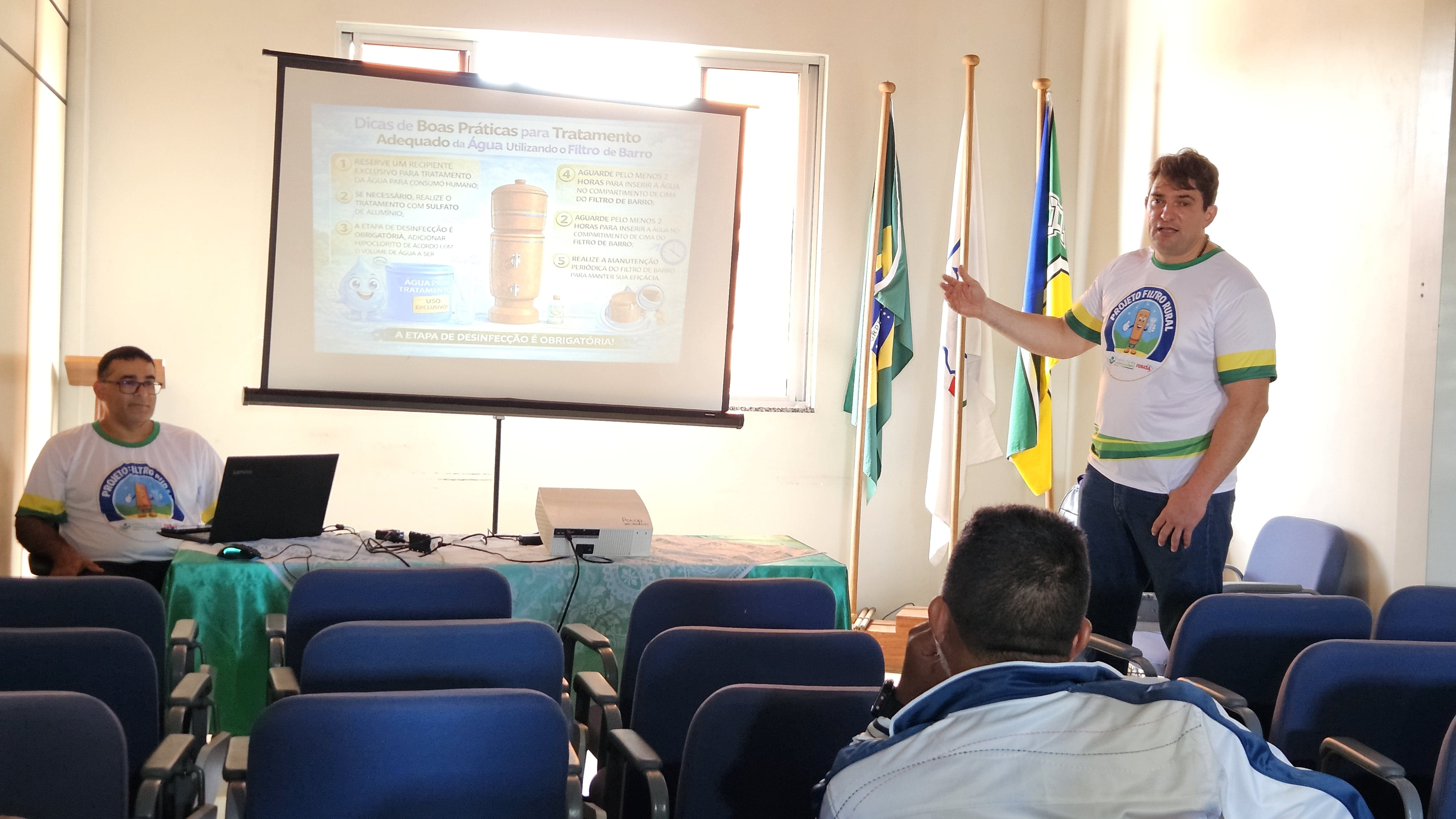 Homem palestrando ao lado de projetor sobre tratamento de água em sala com cadeiras azuis.