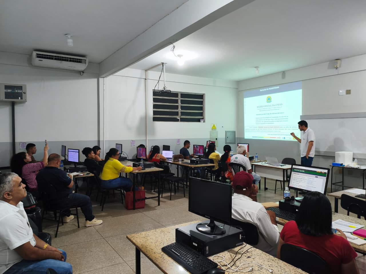 Professor ministra aula teórica para alunos em laboratório de informática com PCs e projetor.