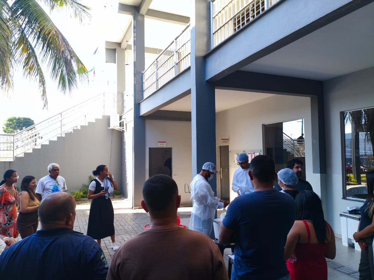 Pessoas de jaleco e touca demonstram coleta de água para grupo em pátio externo de prédio.