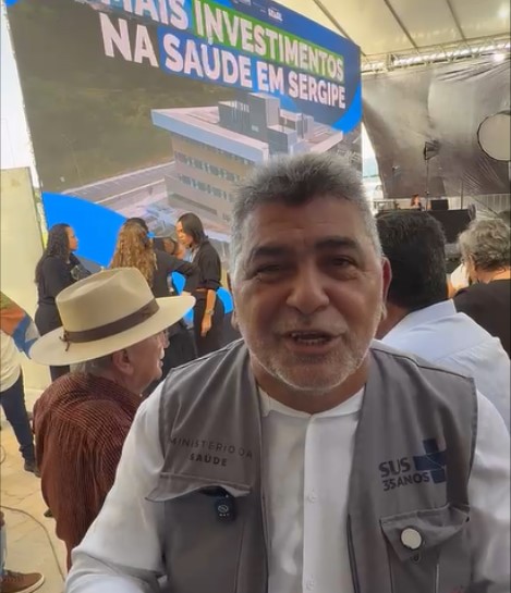 Lenildo Morais, vestindo colete do Ministério da Saúde, fala em primeiro plano com painel de fundo.