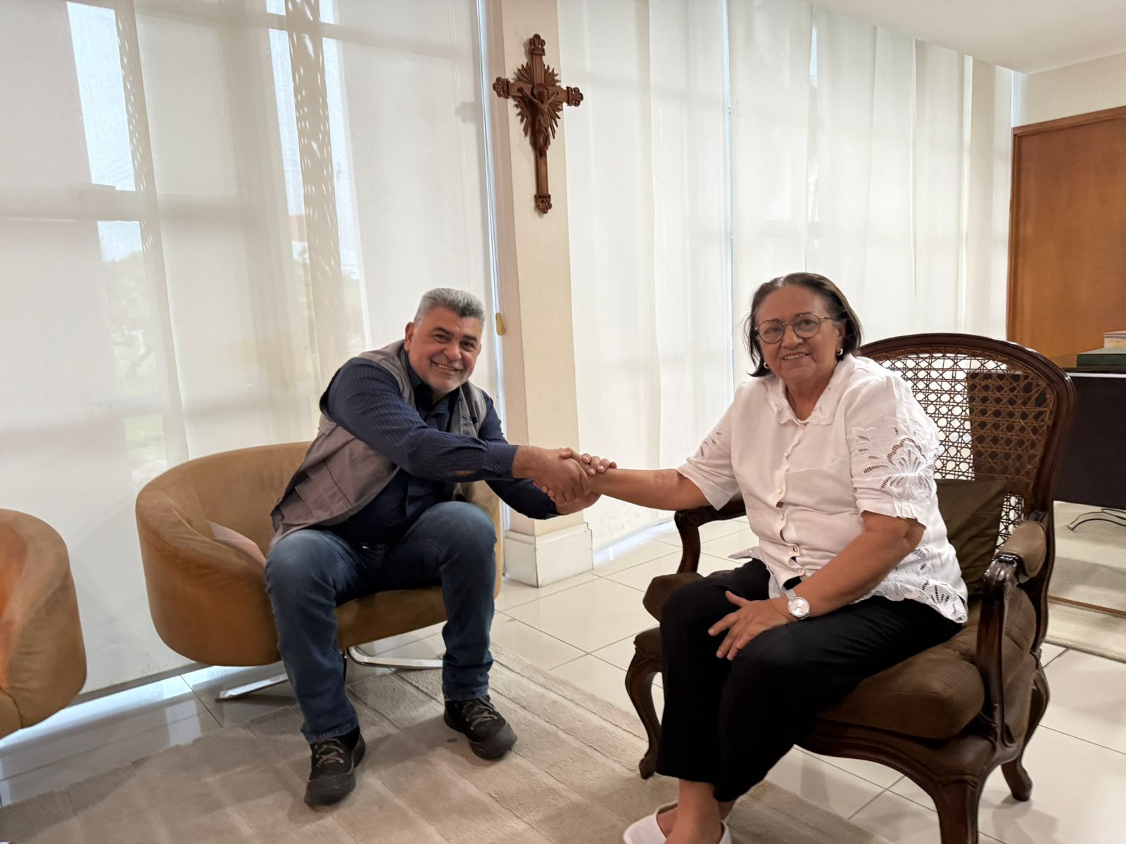 Lenildo Morais e a governadora Fátima Bezerra sorriem e apertam as mãos sentados em um escritório iluminado.