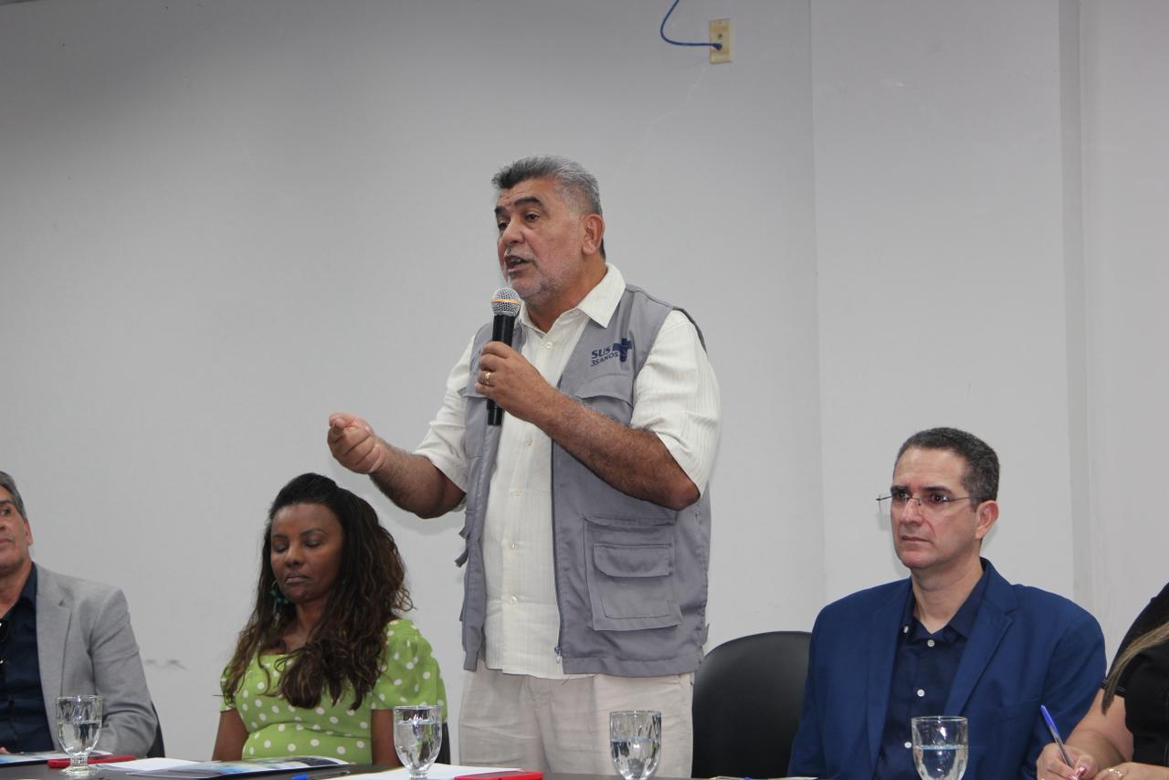 Lenildo Morais, de colete cinza, fala ao microfone em mesa com outros participantes.