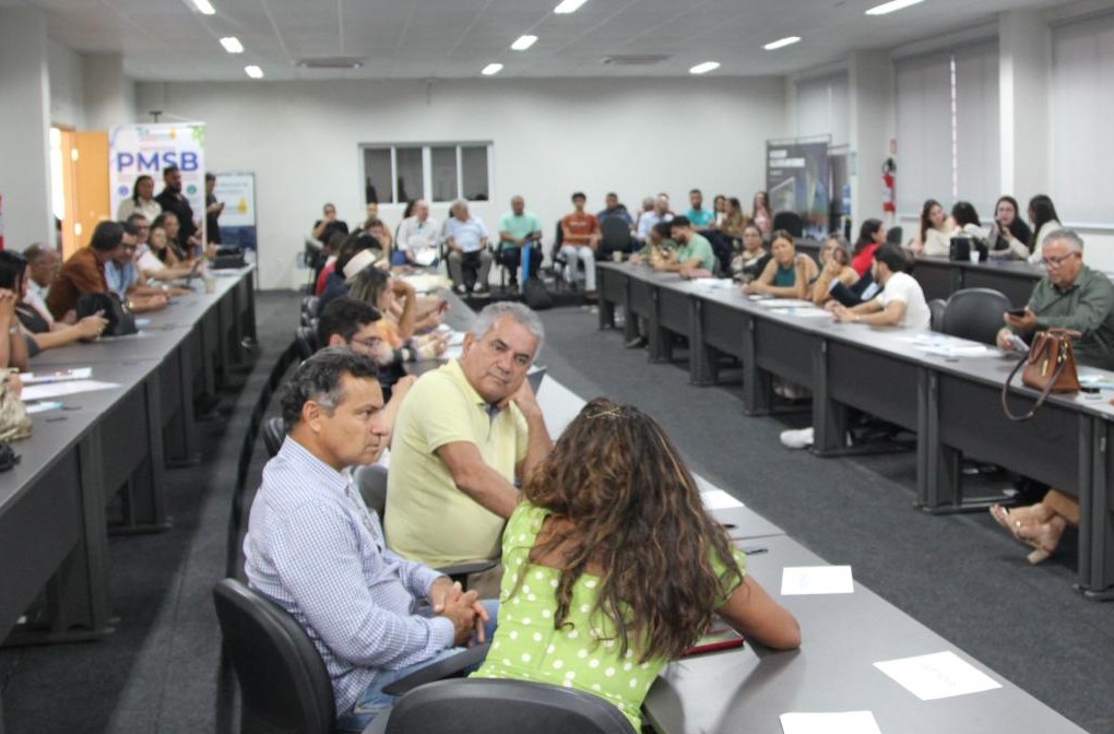 Visão panorâmica de auditório com público assistindo a seminário em mesas em formato de U.