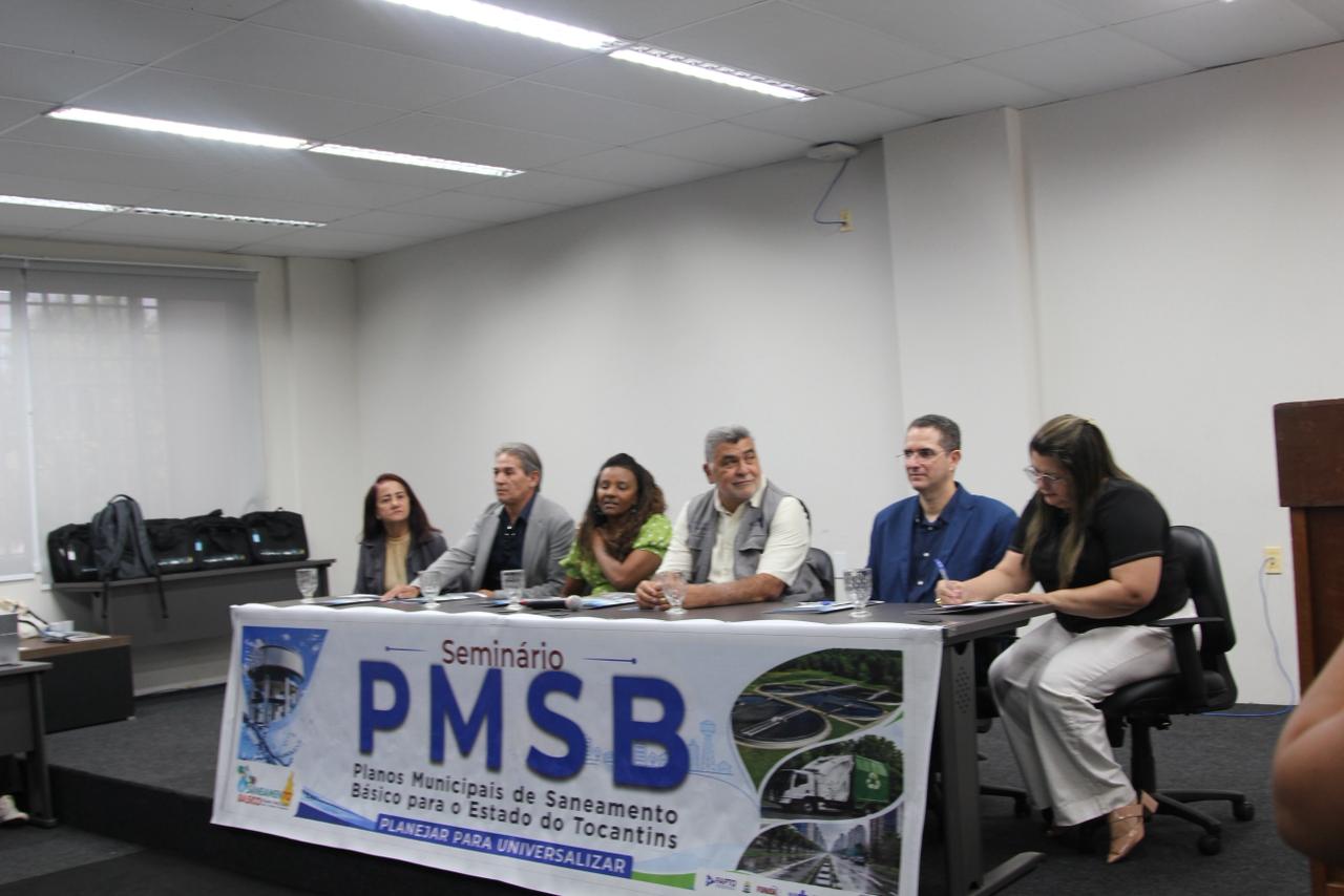 Mesa de palestrantes em seminário com banner frontal escrito "PMSB - Planejar para Universalizar