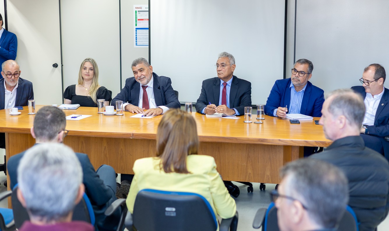 Mesa diretora com seis pessoas sentadas conduzindo reunião institucional para público em primeiro plano.
