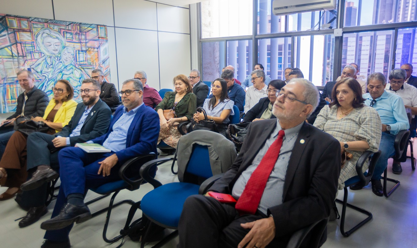 Homem de terno e gravata vermelha sentado em auditório com público assistindo a uma reunião.