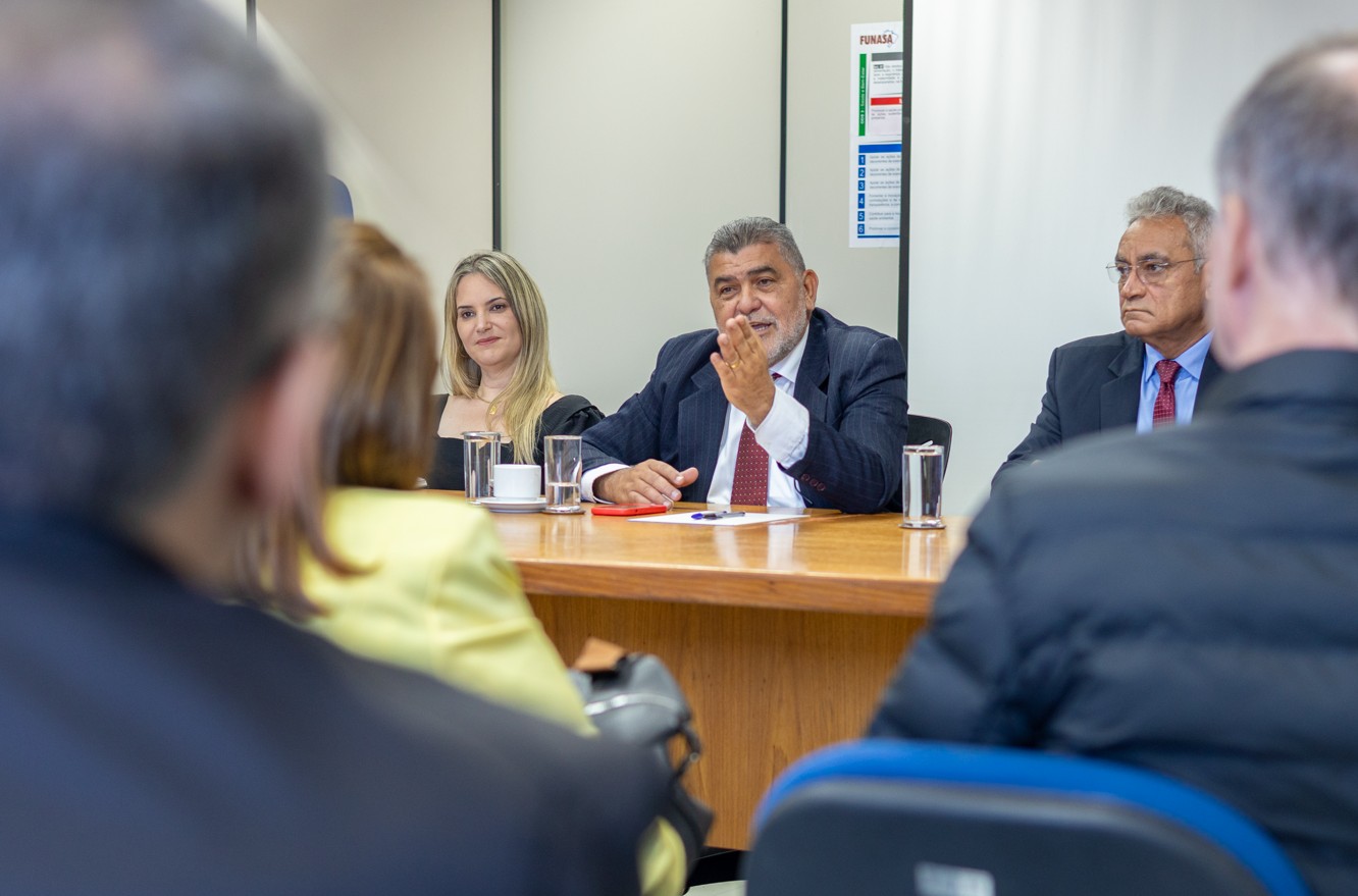 Homem de terno discursa em mesa diretora durante reunião com plateia em primeiro plano.