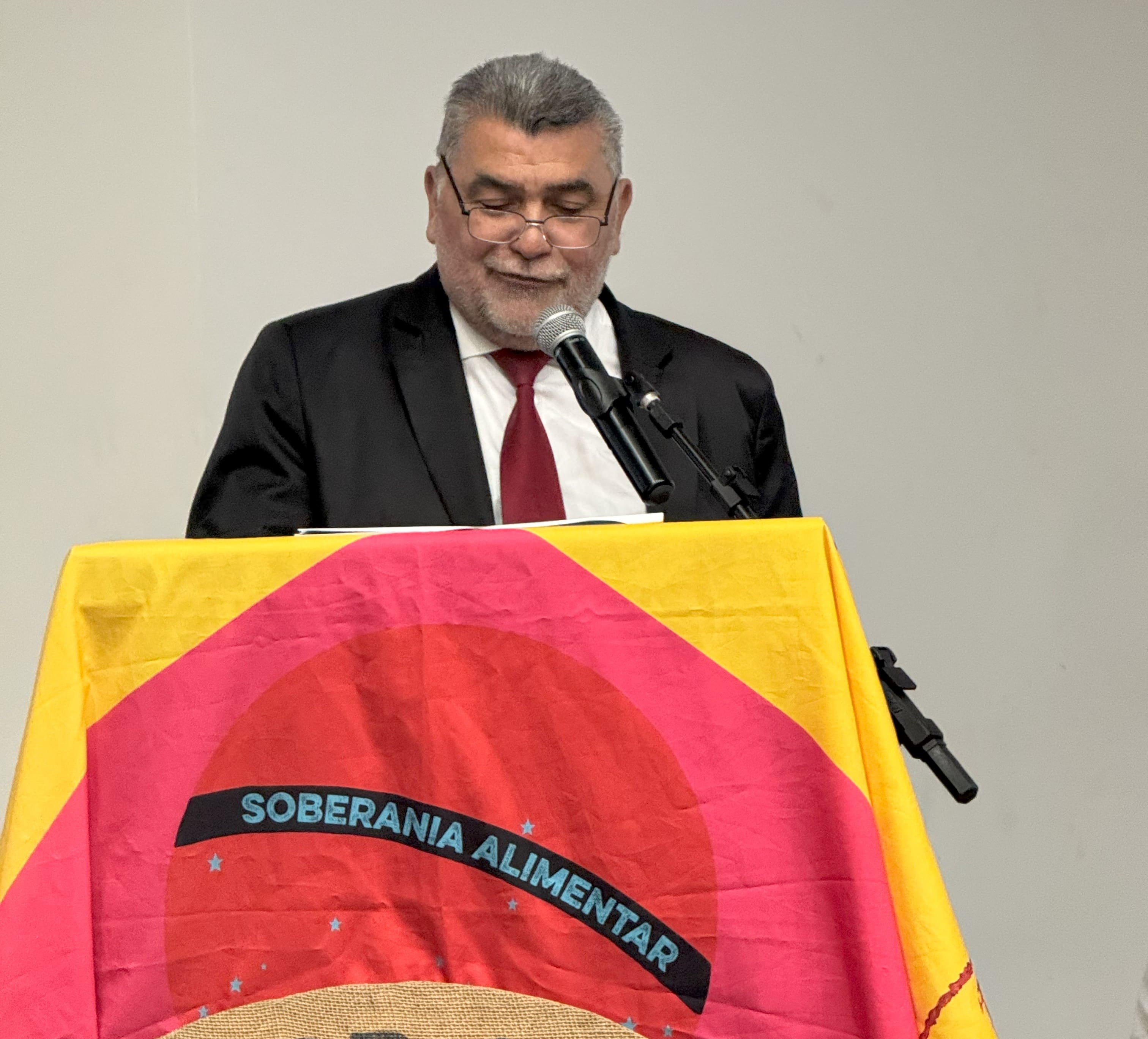 Homem de terno fala ao microfone em púlpito com bandeira onde se lê soberania alimentar.