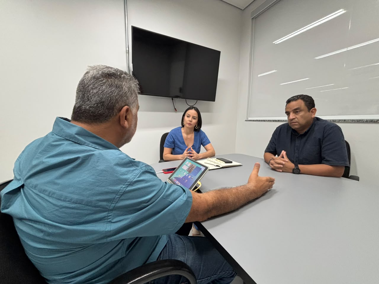 Três pessoas em reunião ao redor de uma mesa cinza em escritório com televisão na parede.