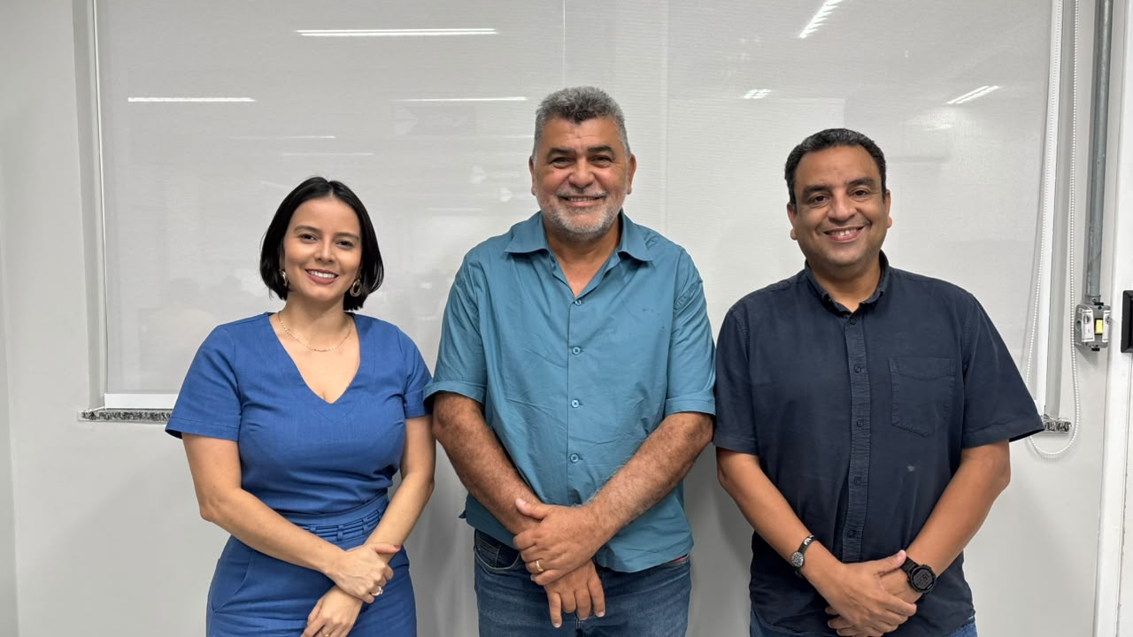 Uma mulher e dois homens sorrindo, lado a lado, em frente a uma parede branca com janela.