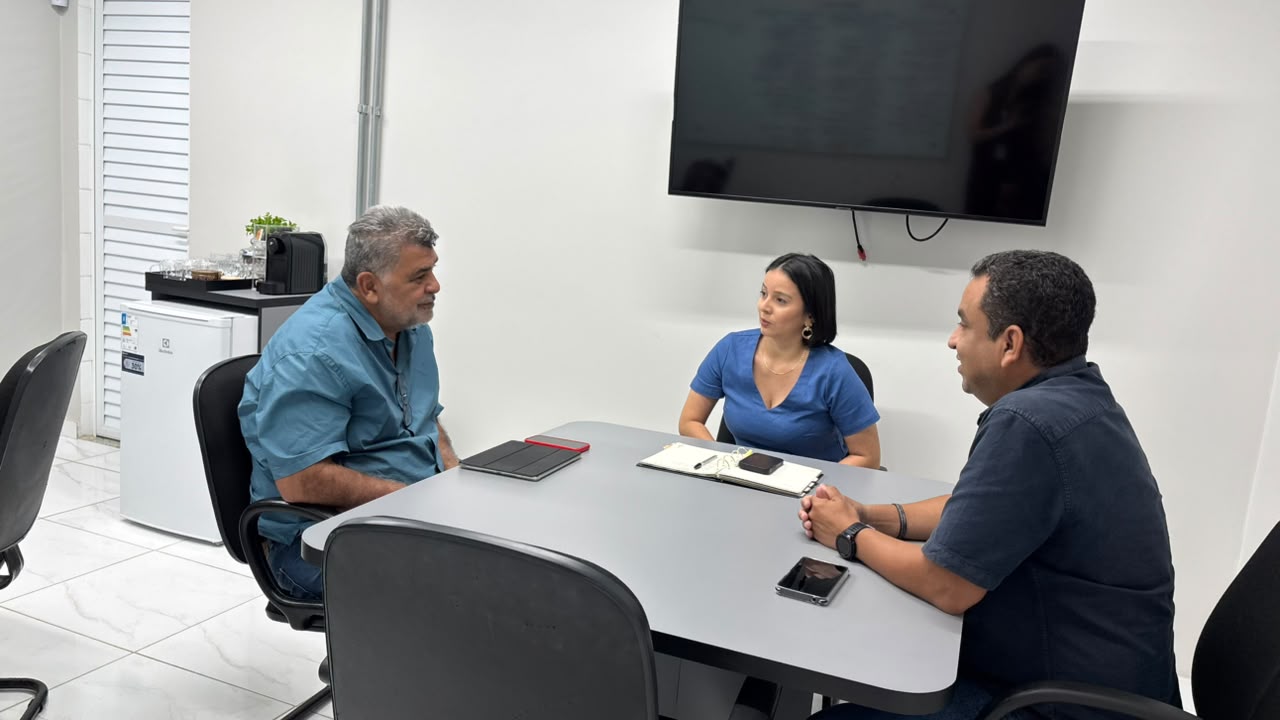 Mulher e dois homens conversando sentados ao redor de uma mesa de reunião em um escritório.