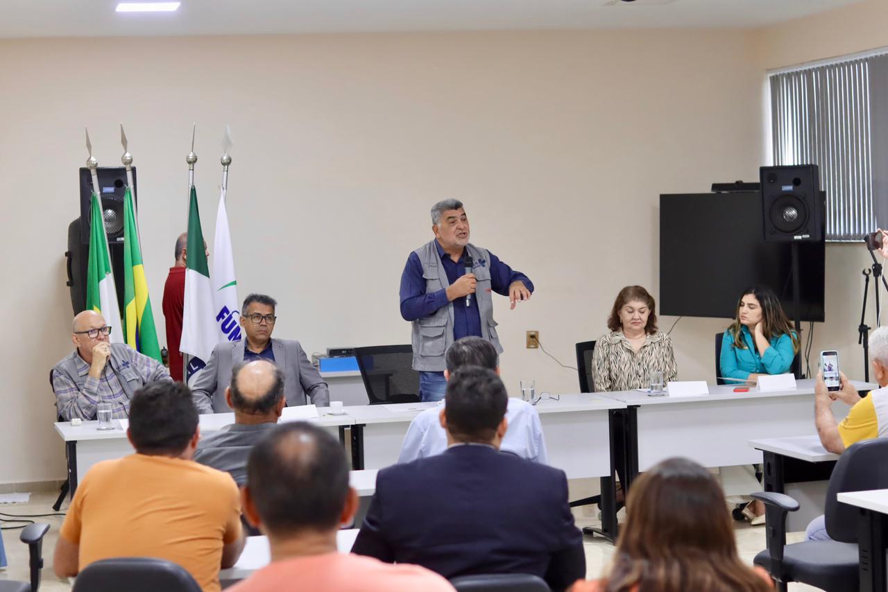 Lenildo Morais discursa ao microfone no centro de mesa diretora para plateia em auditório.