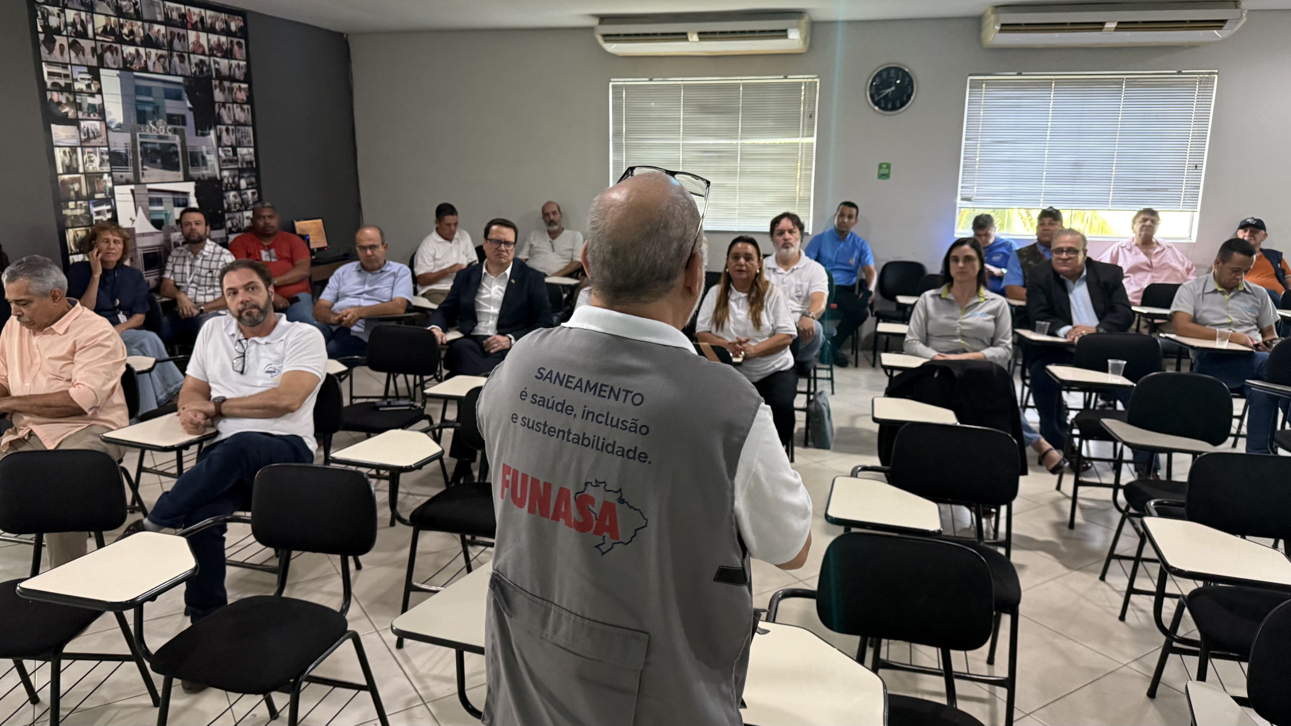Homem de colete cinza da FUNASA fala de costas para uma plateia sentada em sala de reunião.