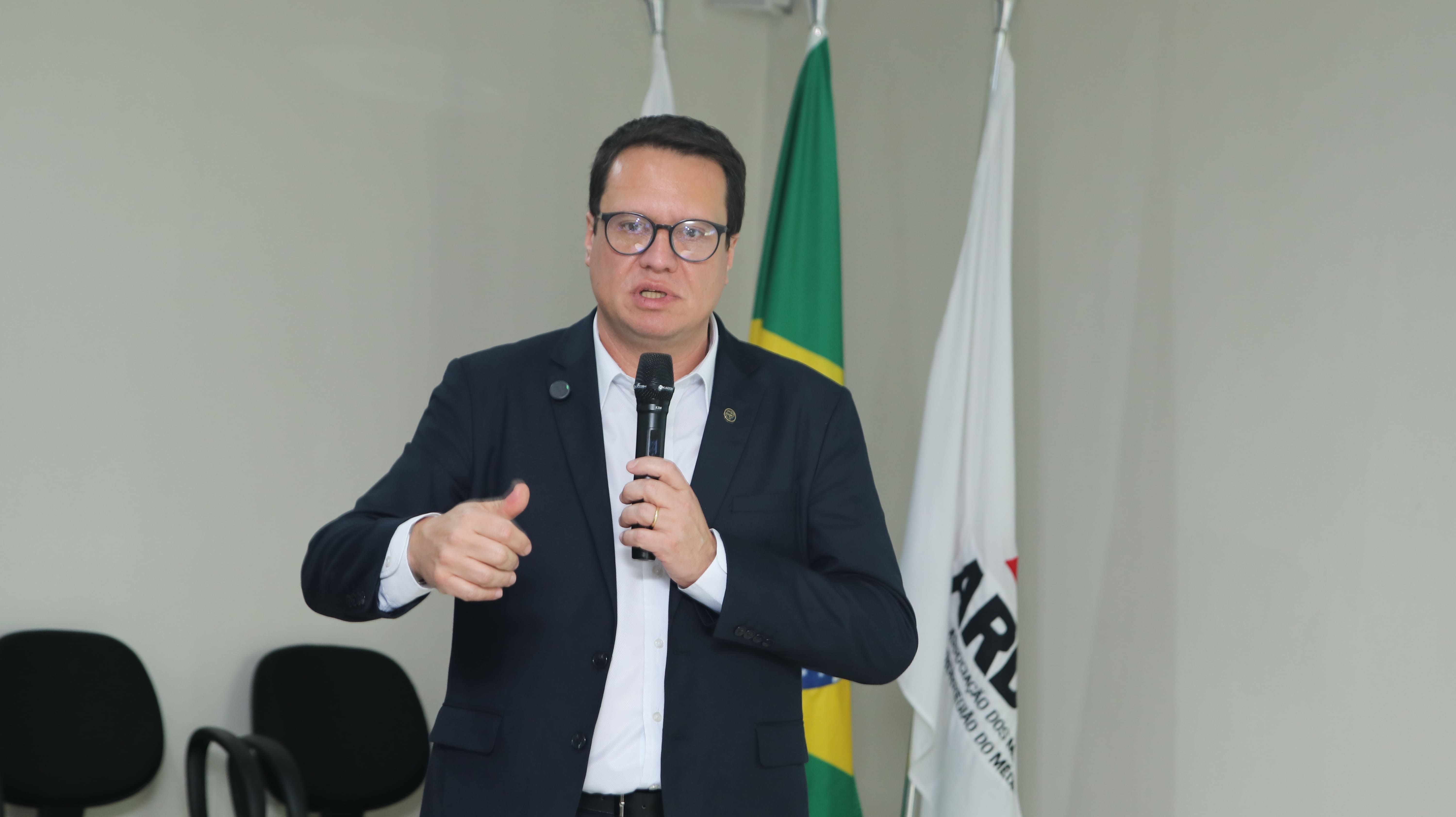 Homem de óculos e terno preto falando ao microfone, com bandeiras do Brasil ao fundo.