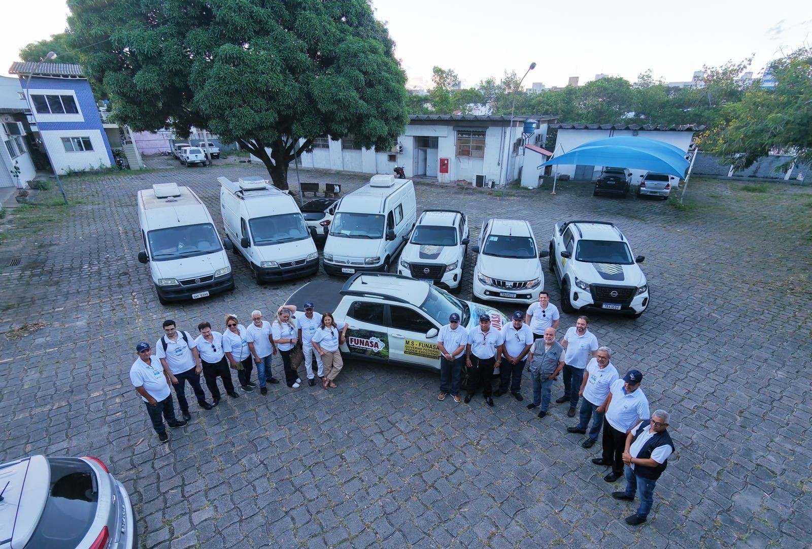 Aérea de grupo uniformizado em pátio de paralelepípedos com vans e picapes da FUNASA.