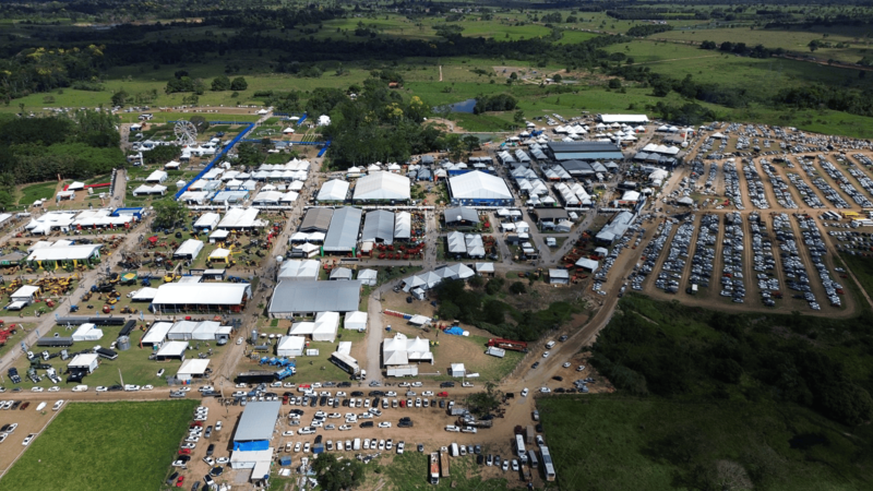 Vista aérea de grande feira agrícola com pavilhões brancos, estacionamentos lotados e ao fundo.