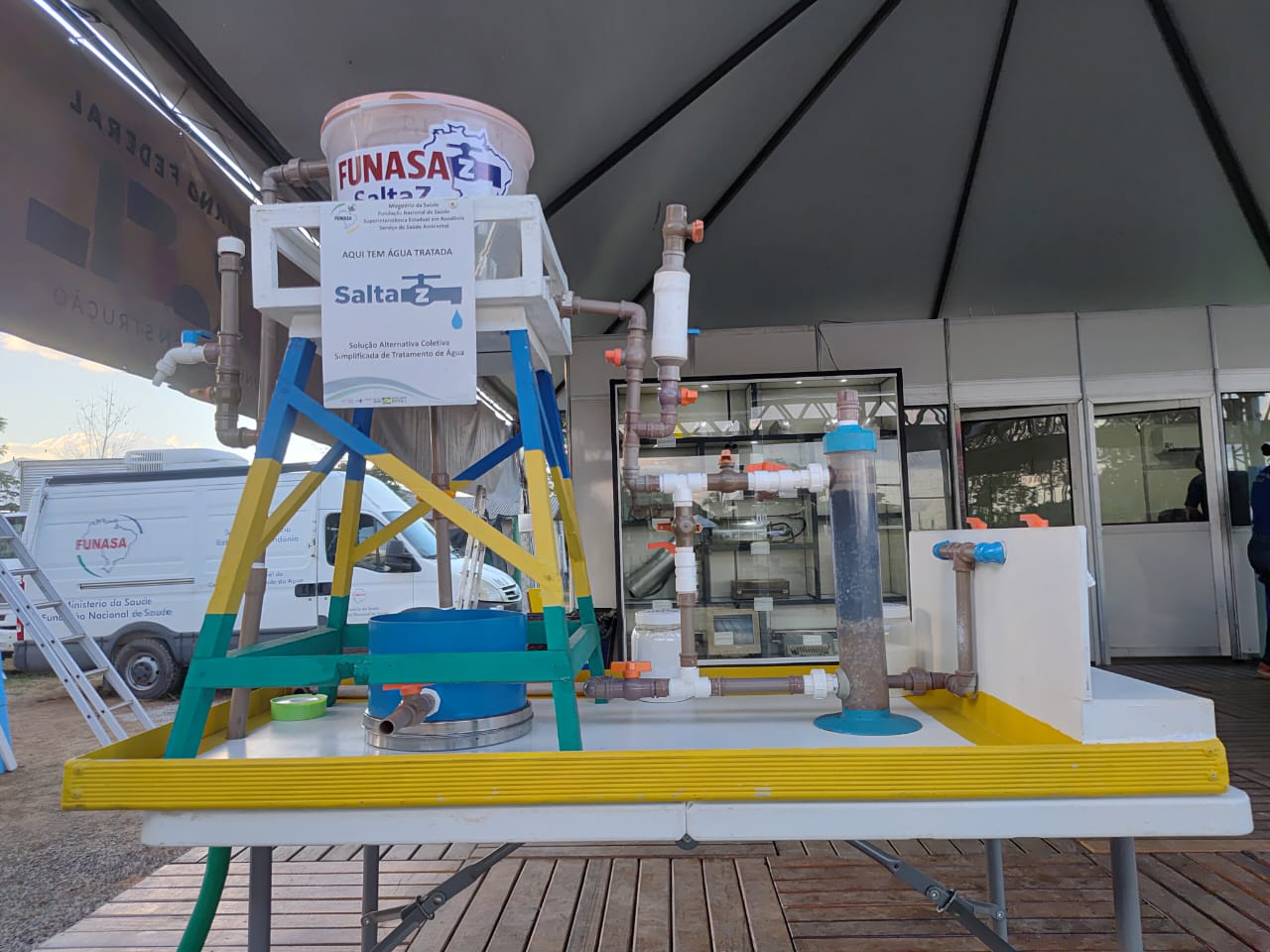 Maquete de sistema de tratamento de água Salta-Z sobre mesa amarela sob tenda em feira de eventos.