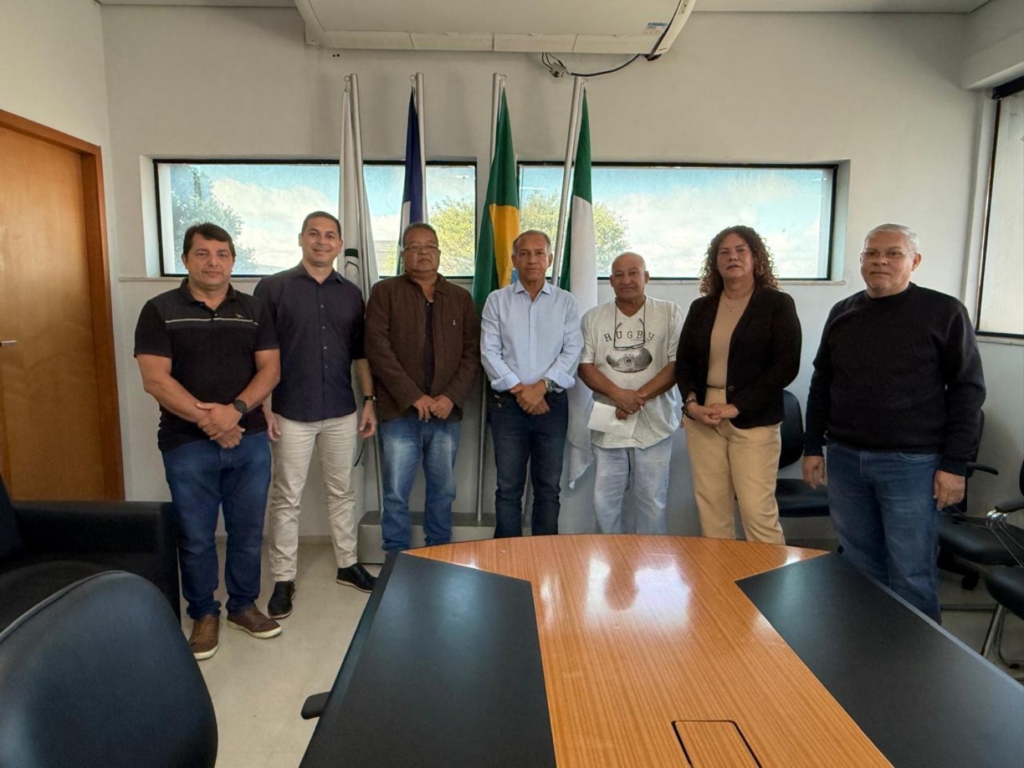 Sete pessoas posam alinhadas em uma sala de reuniões, à frente de bandeiras e de uma mesa de madeira.