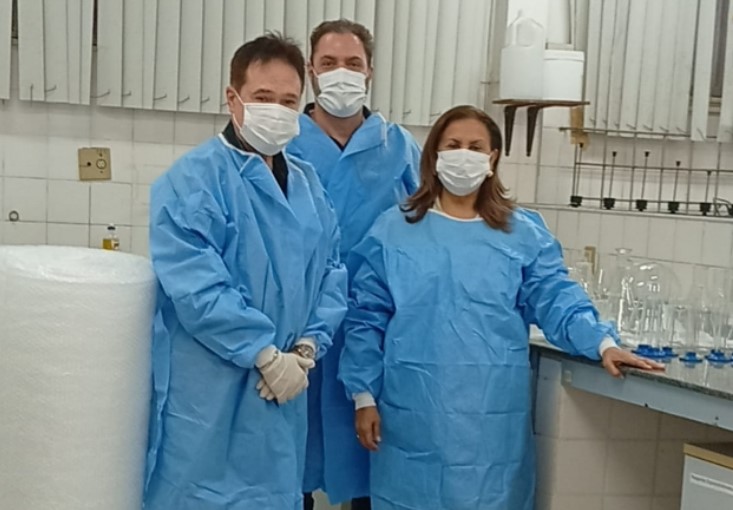 Três profissionais de avental azul e máscara posam em laboratório com vidrarias ao fundo.