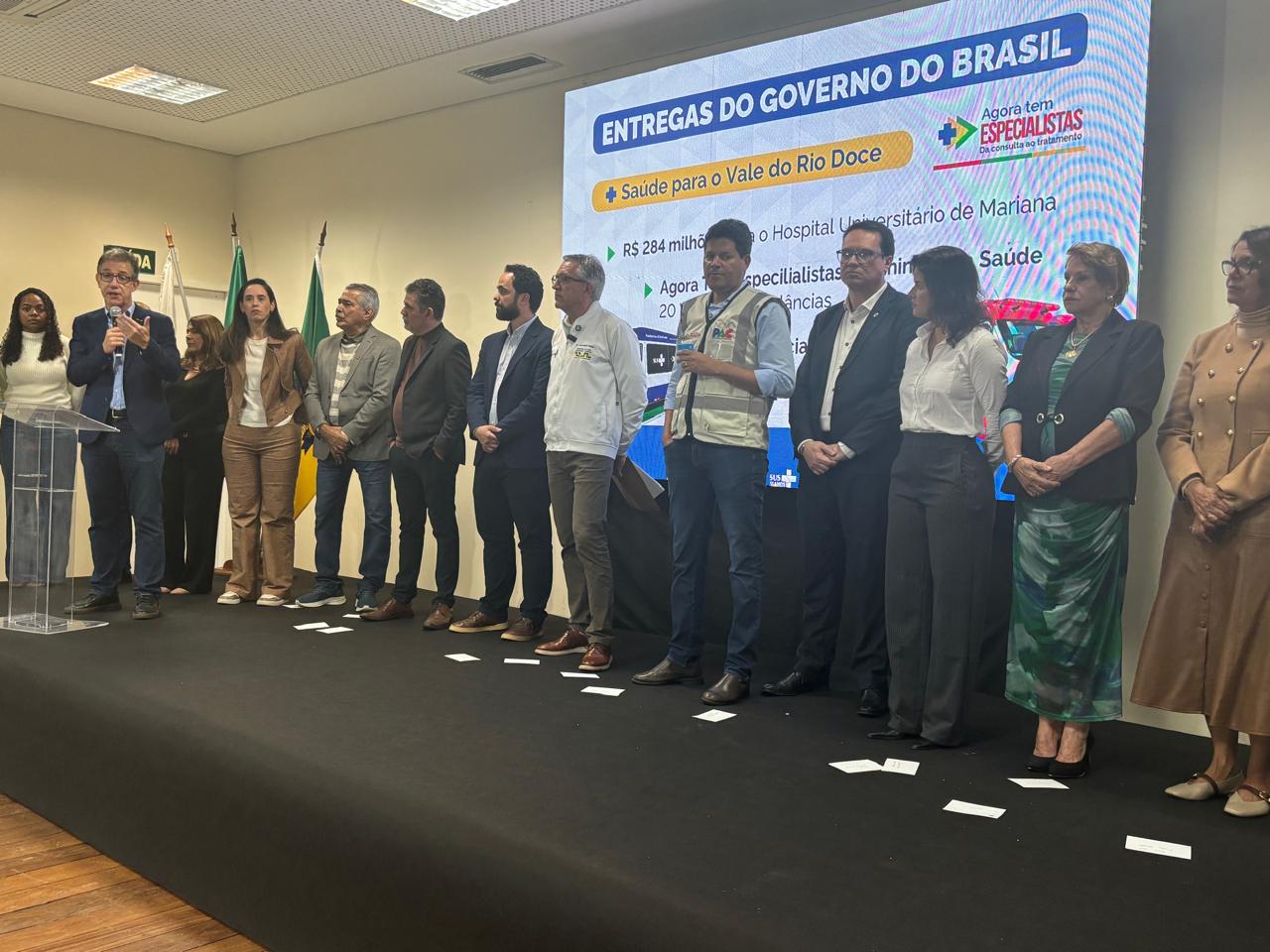 Grupo de autoridades enfileiradas em palco diante de telão sobre entregas do governo na saúde.