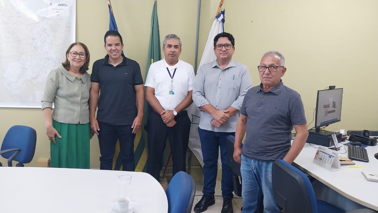 Uma mulher e quatro homens posam alinhados em um escritório em frente a bandeiras.