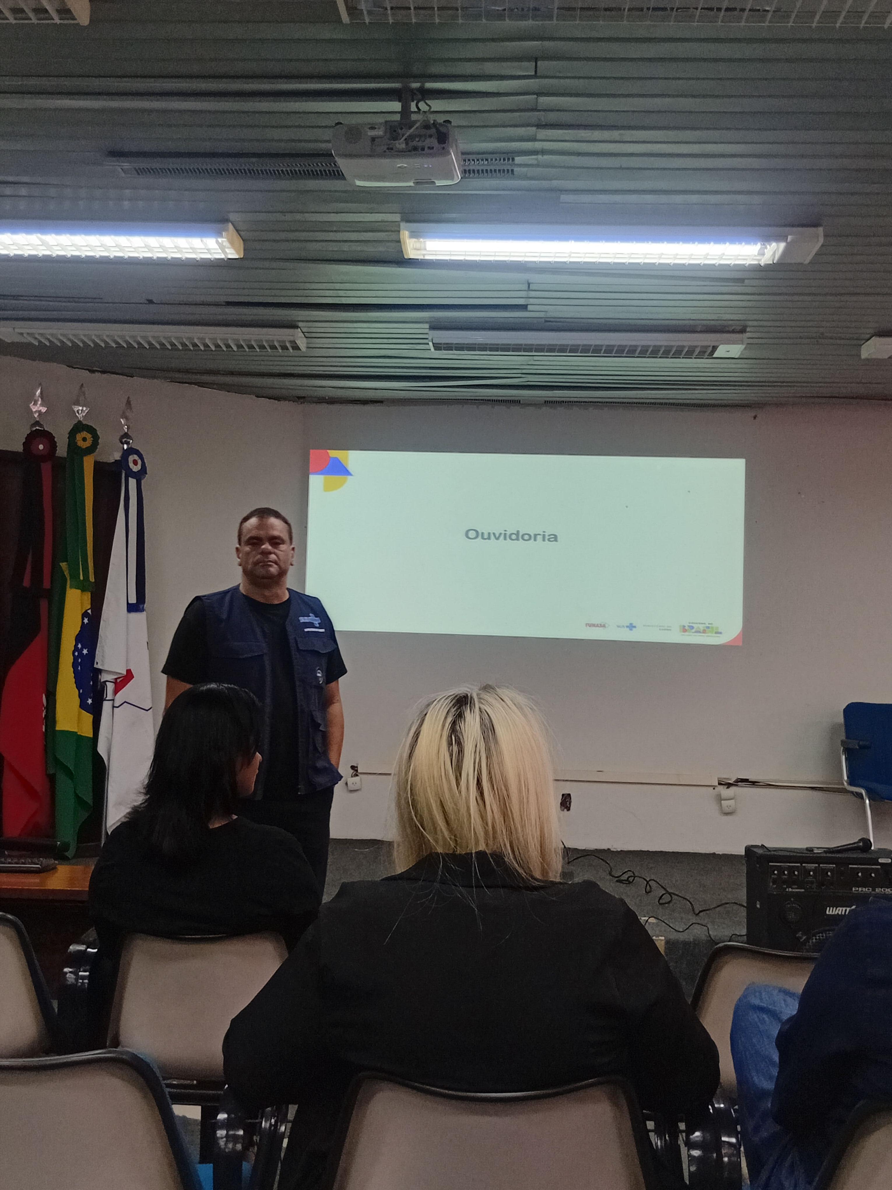 Homem em colete azul palestra diante de tela com a palavra 'Ouvidoria' para plateia sentada.