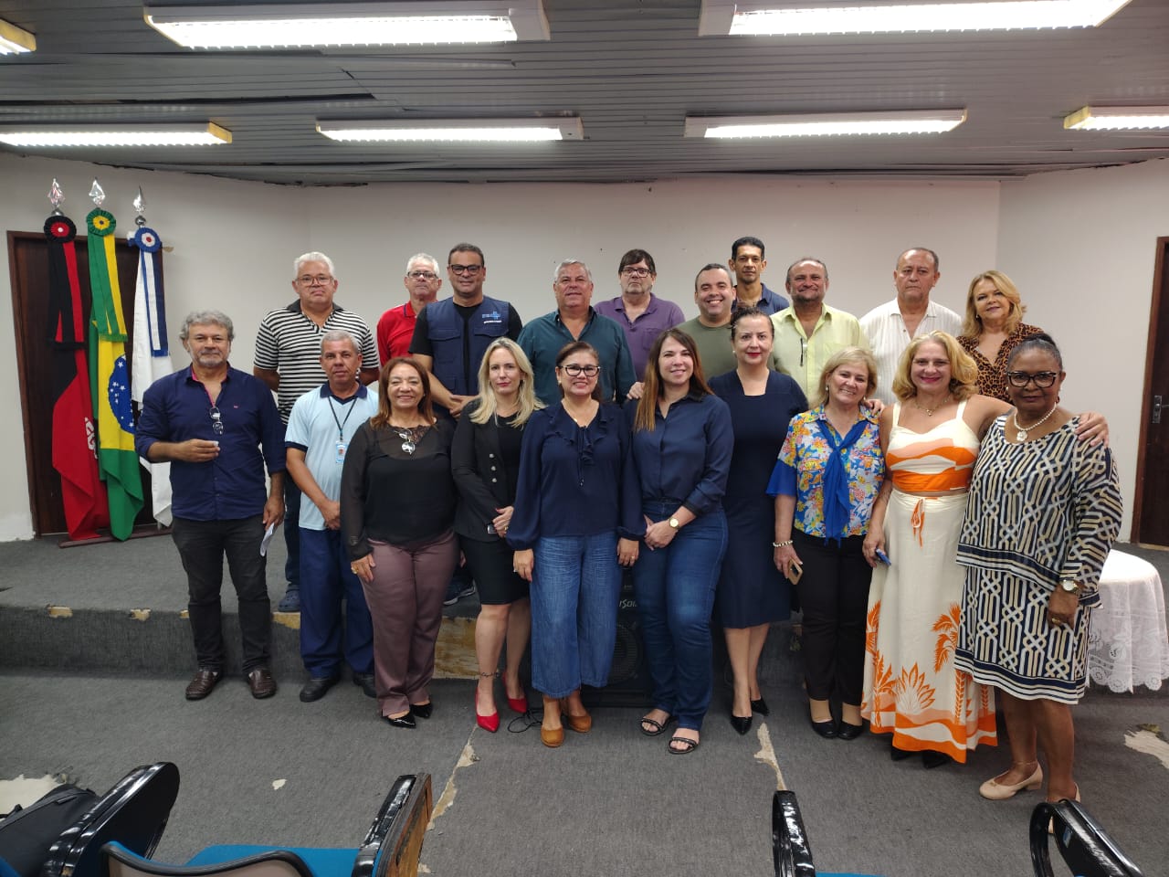 Grupo de vinte pessoas posa para foto oficial em frente a bandeiras em um auditório.