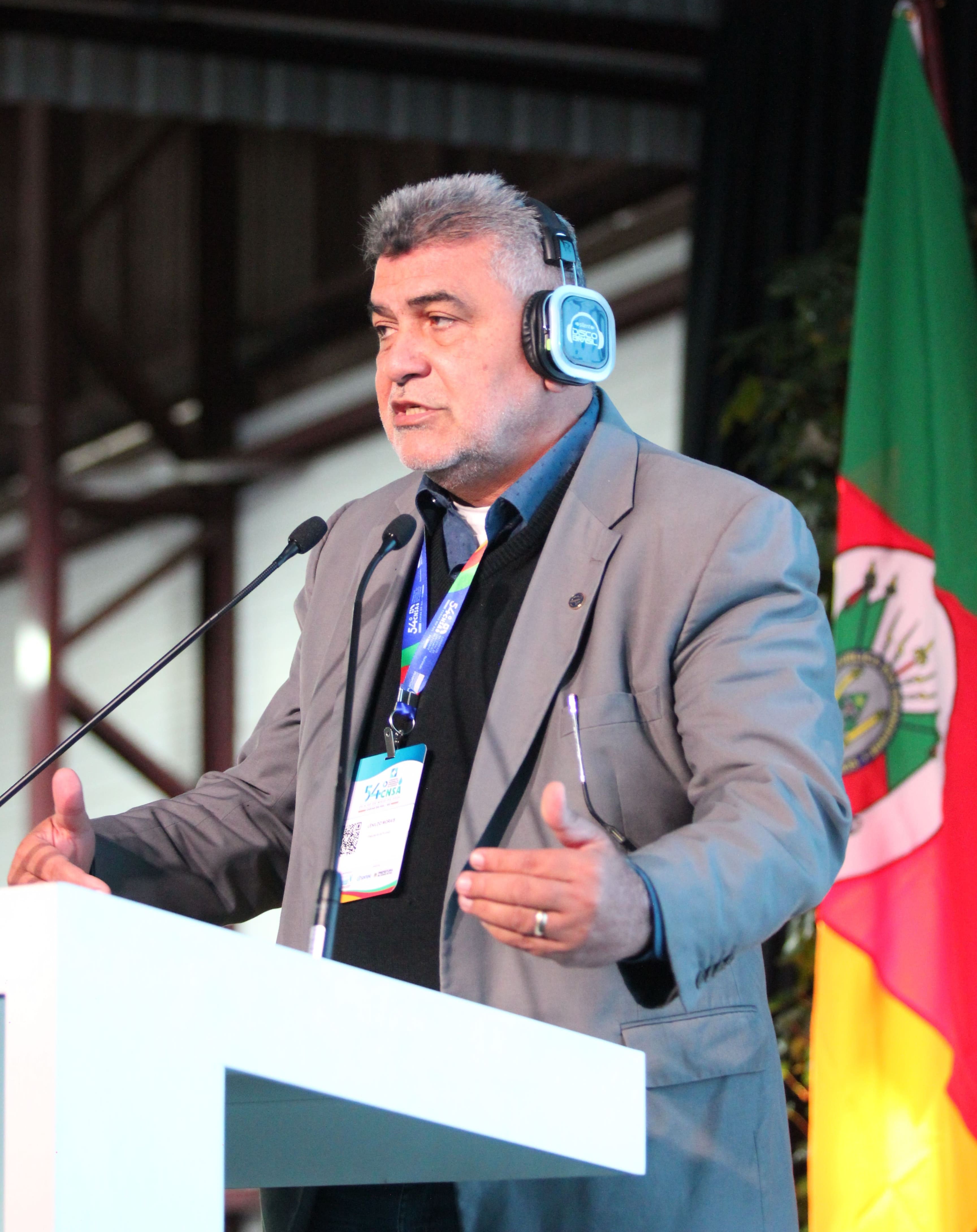 Plano médio de Lenildo Morais gesticulando no púlpito, com bandeira ao fundo.