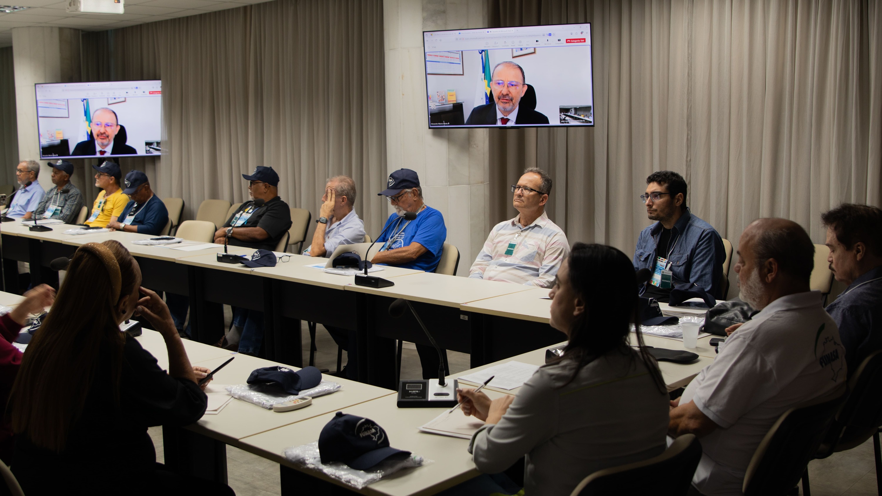 Pessoas em reunião ao redor de mesas brancas, assistindo a um homem em videochamada nas telas da sala.