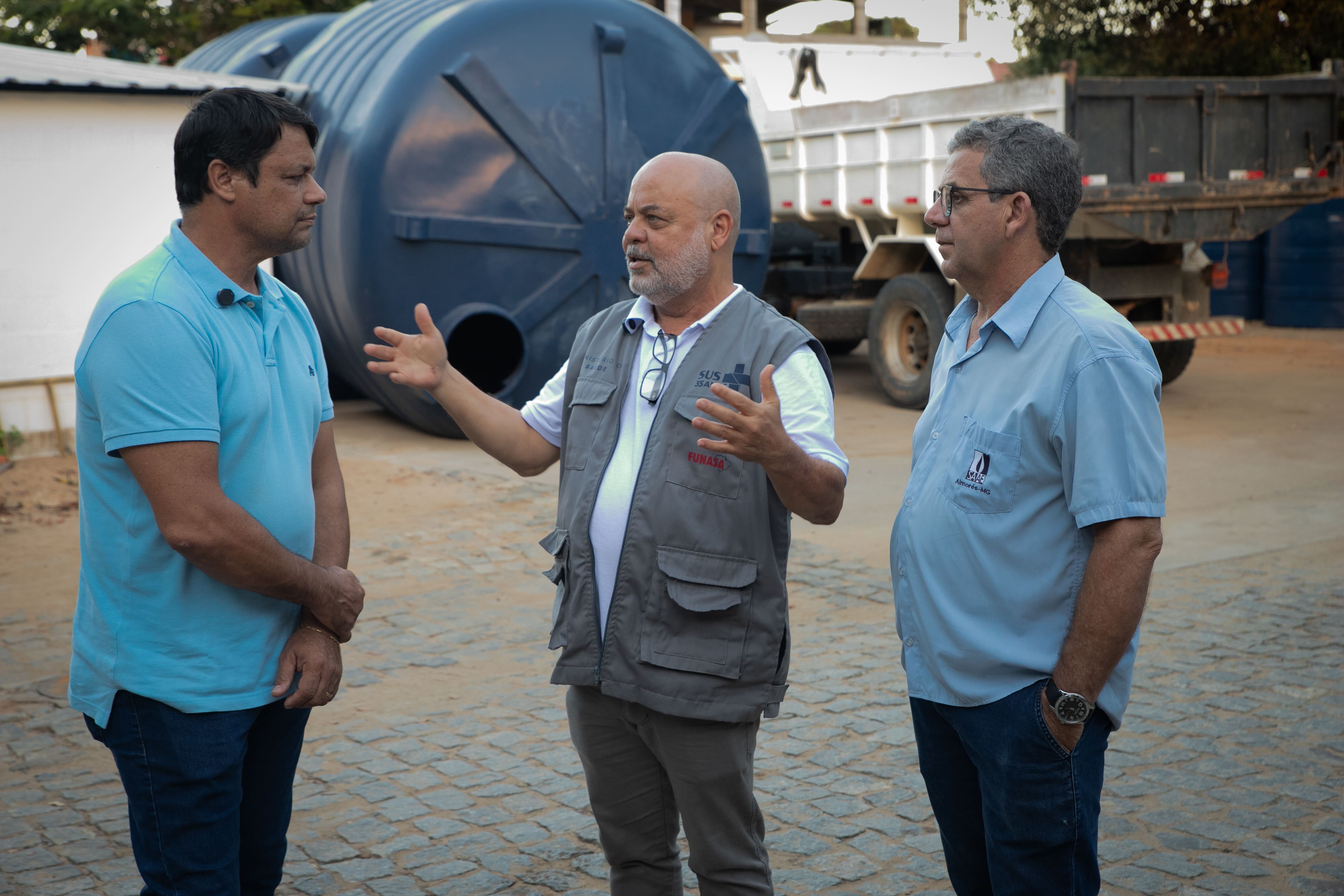 Três homens conversam em pátio externo ao lado de grande reservatório azul e caminhão.