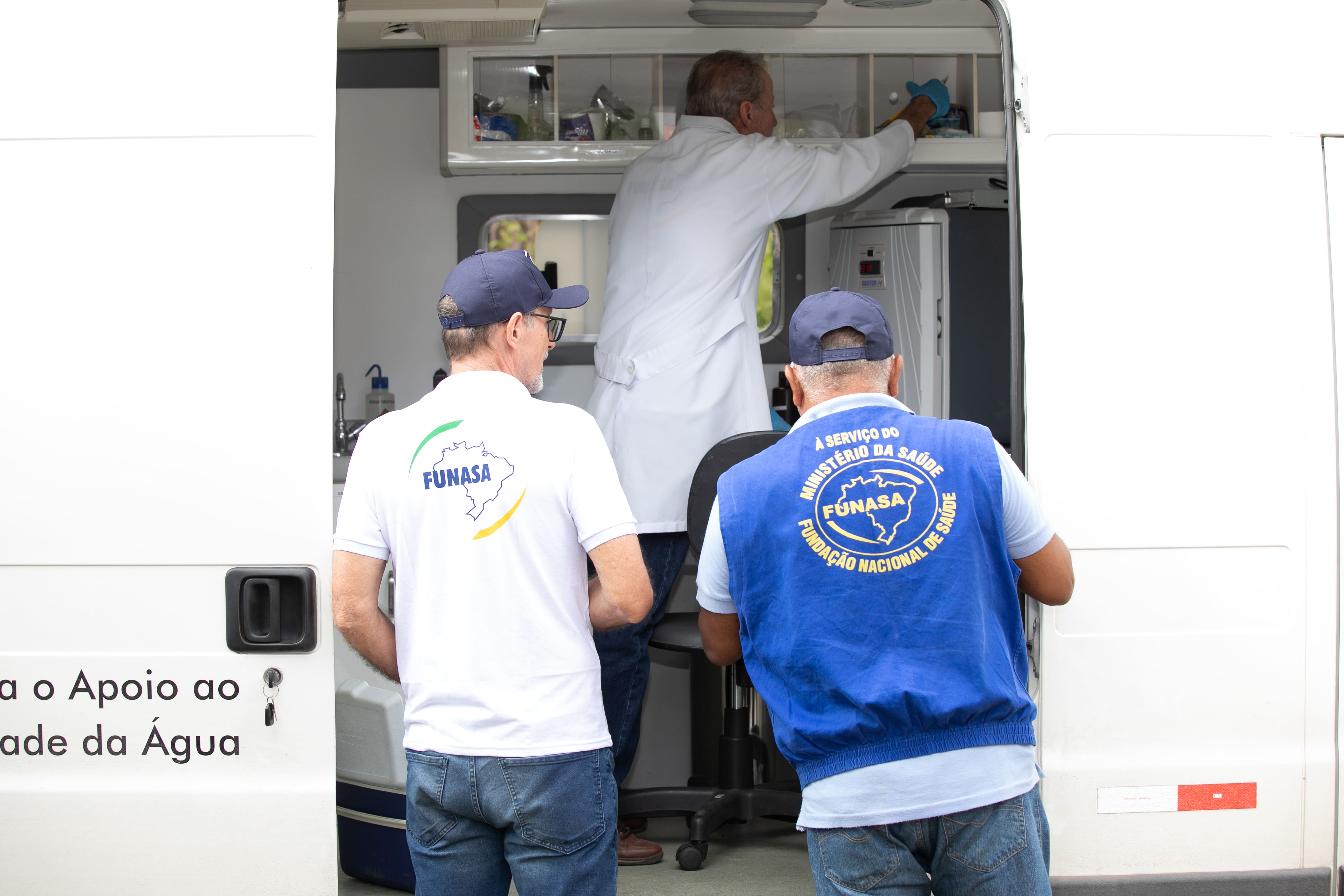 Dois técnicos uniformizados observam colega trabalhando dentro de veículo de laboratório.