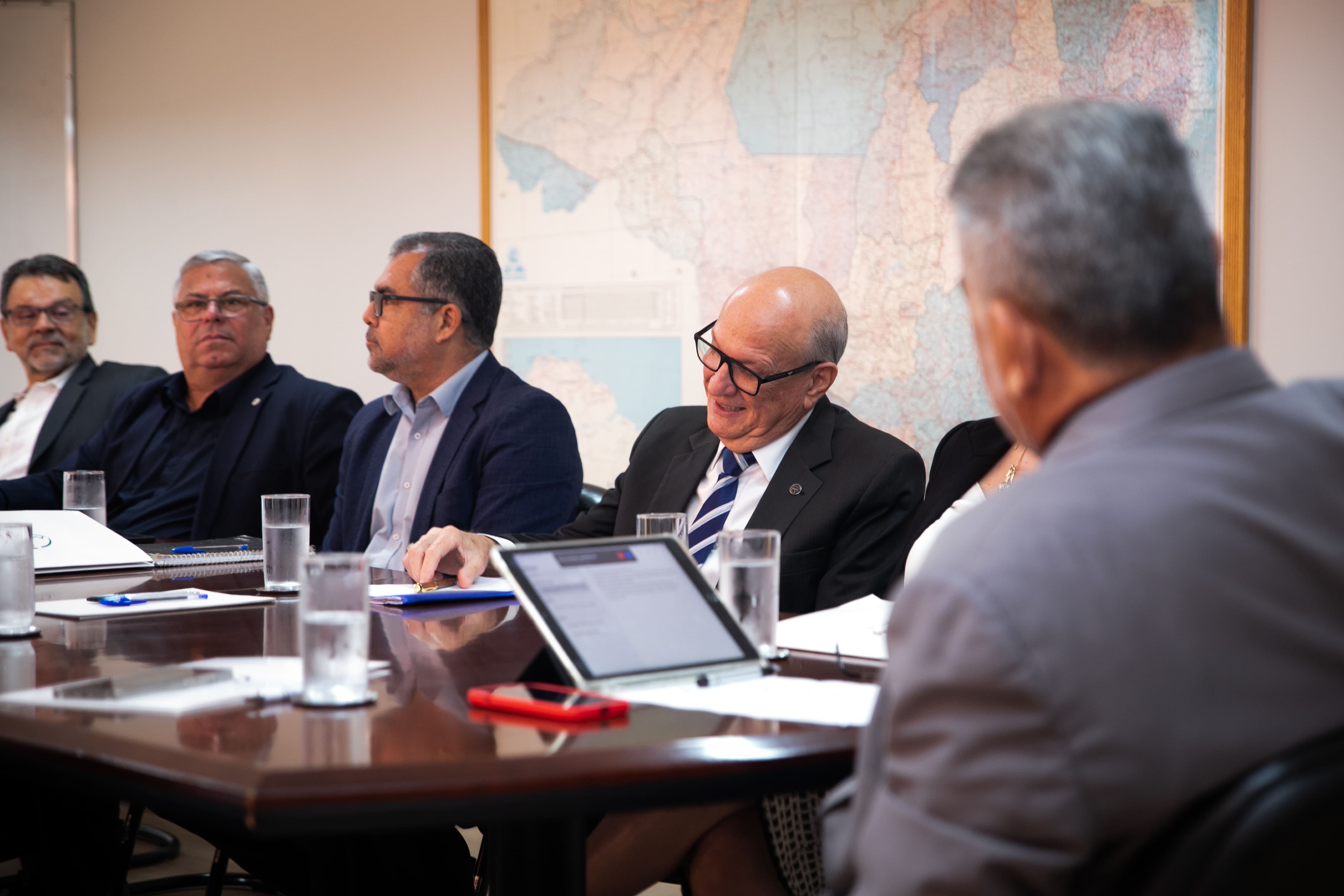 Participantes de reunião conversam em mesa com mapa do Brasil ao fundo.