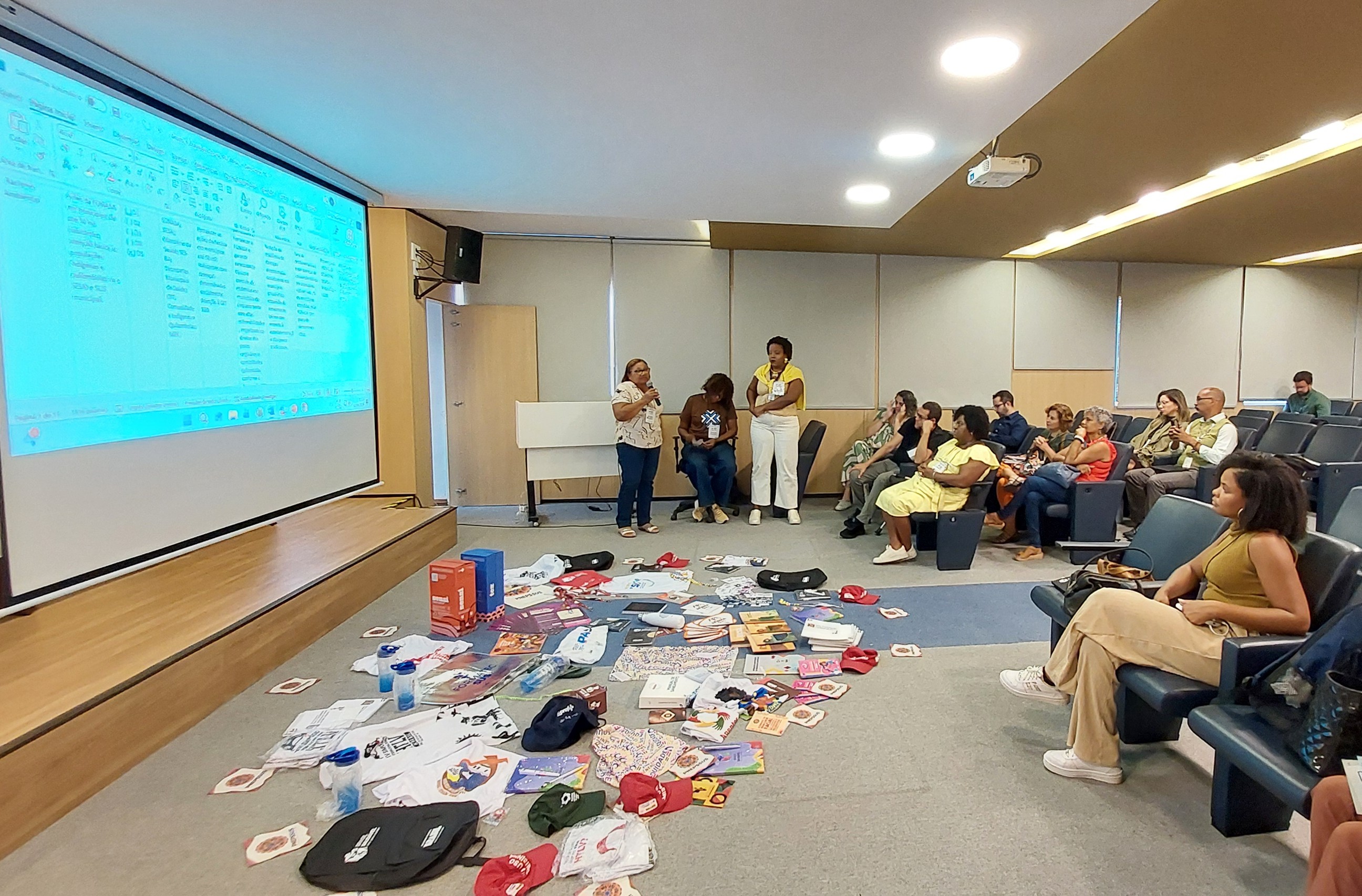 Mulher fala ao microfone em auditório para público sentado, ao lado de projeção em tela branca.