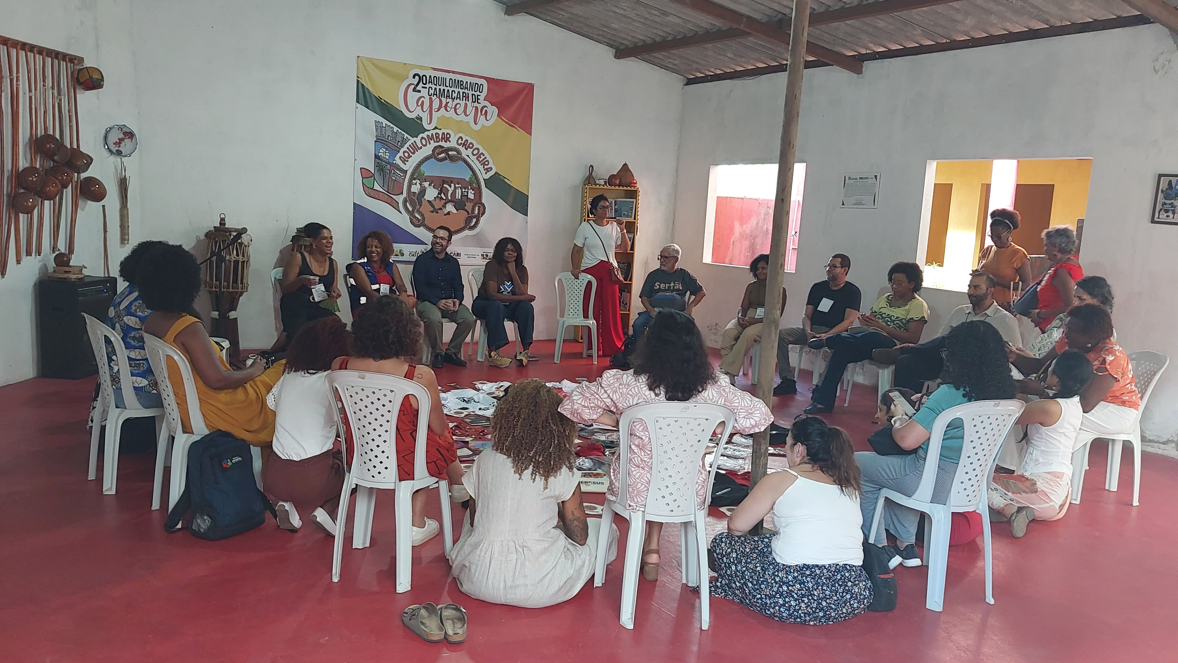 Grupo em círculo sentado em sala com banner de capoeira ao fundo e materiais espalhados no chão.