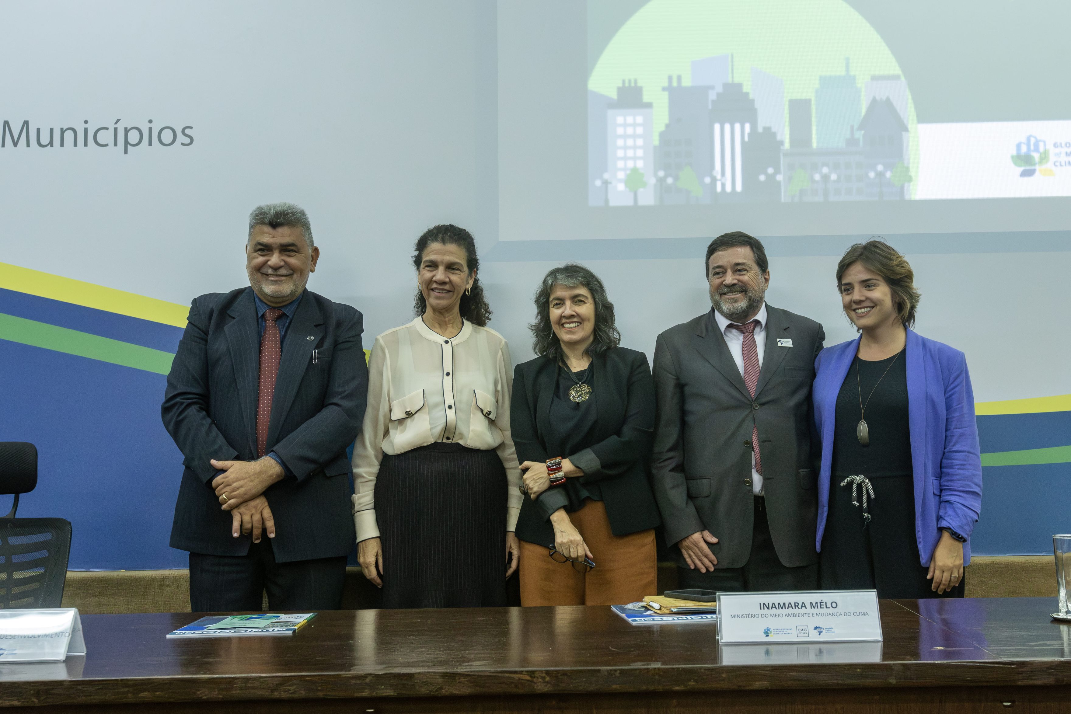 Cinco pessoas em pé, homens e mulheres, lado a lado atrás de uma mesa, posando para foto.