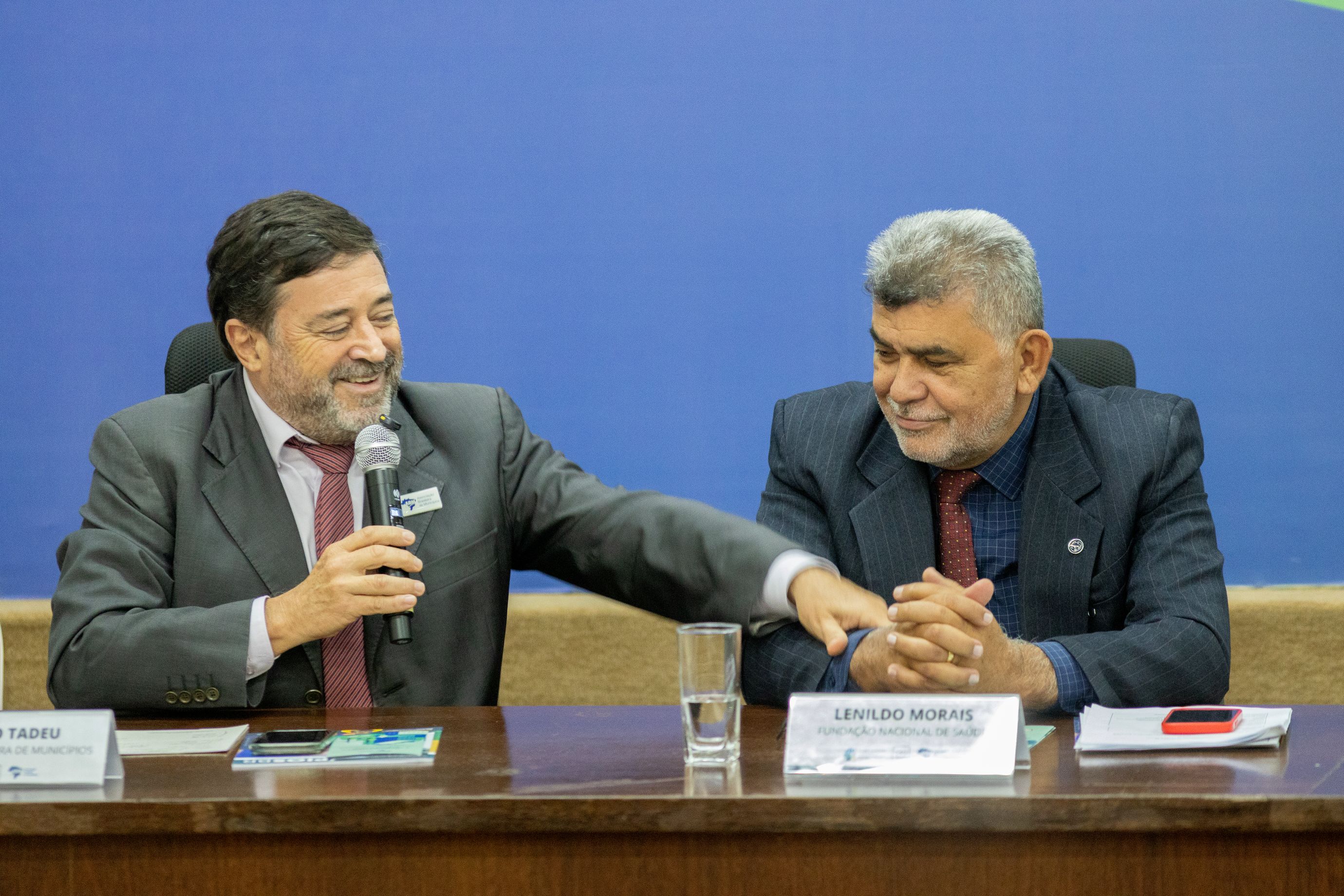 Dois homens de terno cinza, um moreno fala algo ao microfone, o outro grisalho escuta com atenção.