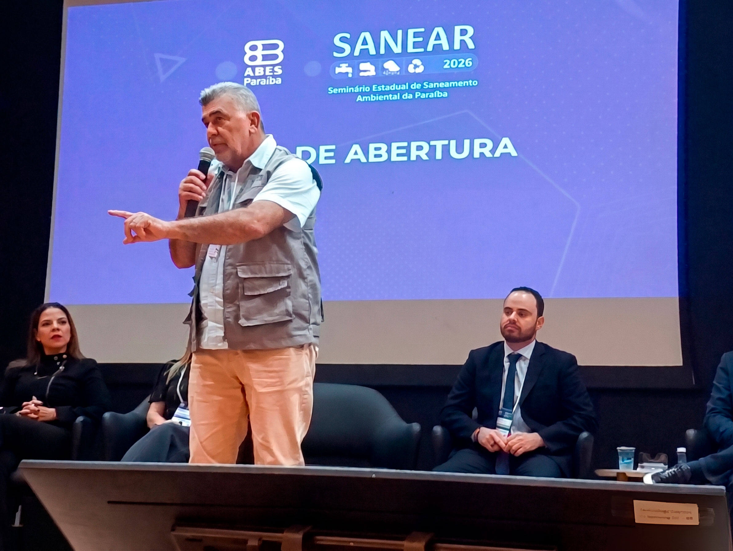 Homem em palco, palestrando em evento chamado Sanear.