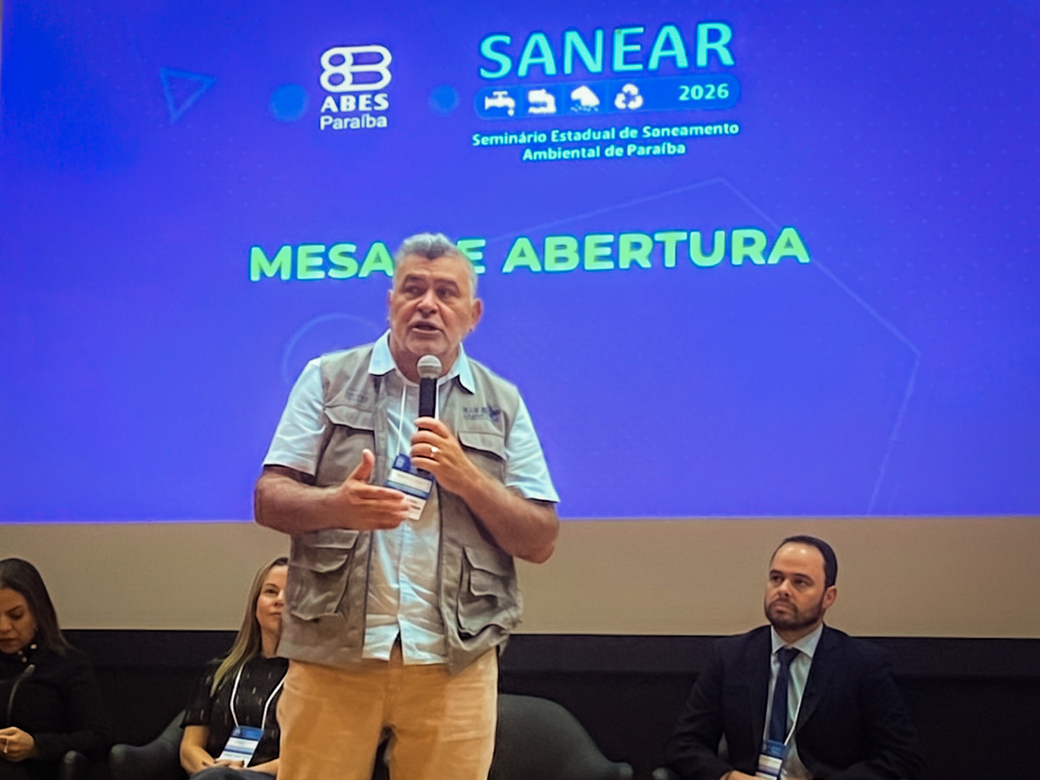 Homem em palco, palestrando em evento chamado Sanear.