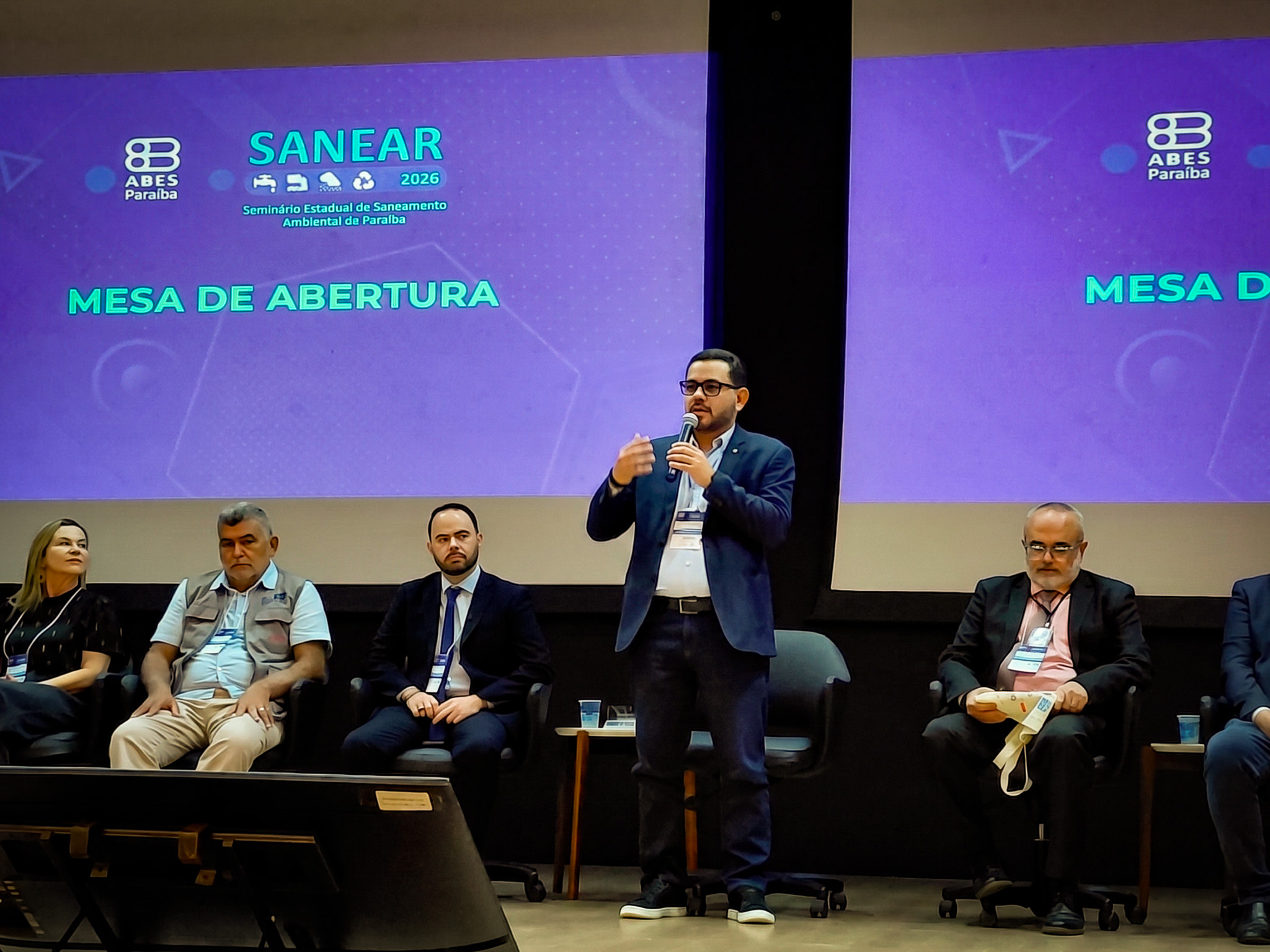 Homem de terno azul palestrando em palco de evento com pessoas assistindo.