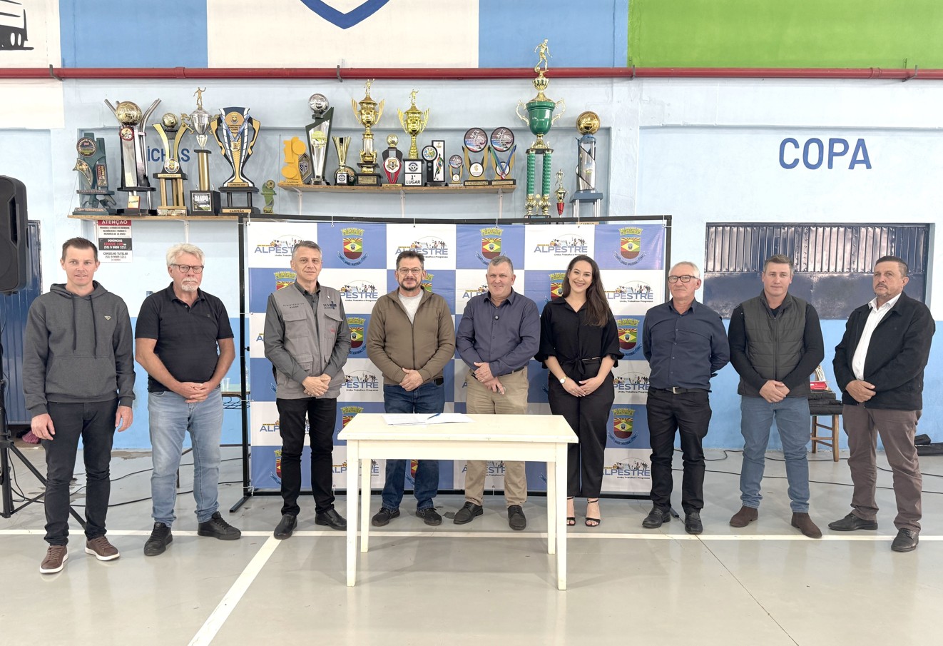 Nove pessoas posam alinhadas atrás de mesa branca e painel azul e branco da prefeitura de Alpestre.