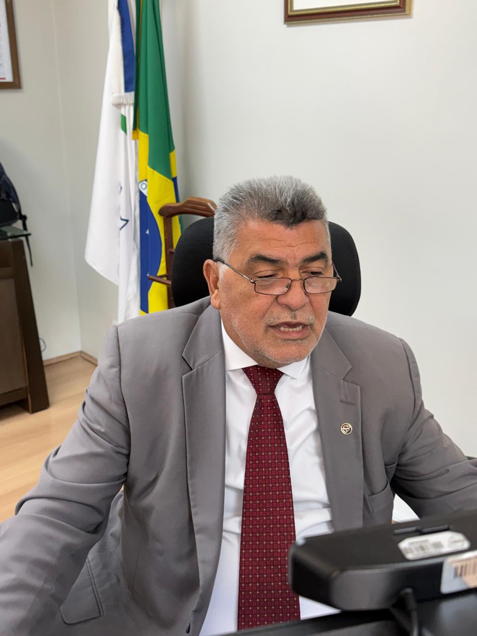 Homem de terno cinza e óculos fala sentado em escritório com bandeiras ao fundo.