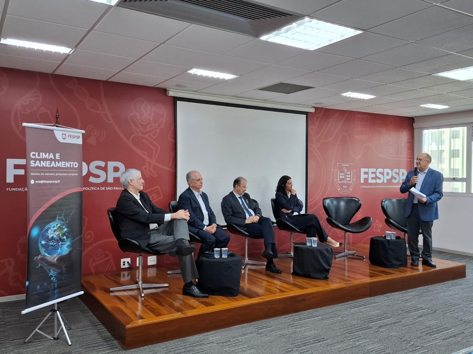 Cinco palestrantes em um palco da FESPSP sobre Clima e Saneamento; um homem fala ao microfone.